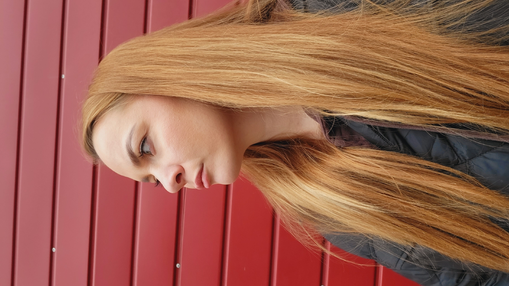 A young woman with long, light brown hair is seen in profile, facing left. Her skin is fair and her eyes are light colored. She is wearing a dark quilted jacket. The background is a red metal wall with vertical lines, possibly a building facade. The photograph was taken in Chaikovsky, Russia.