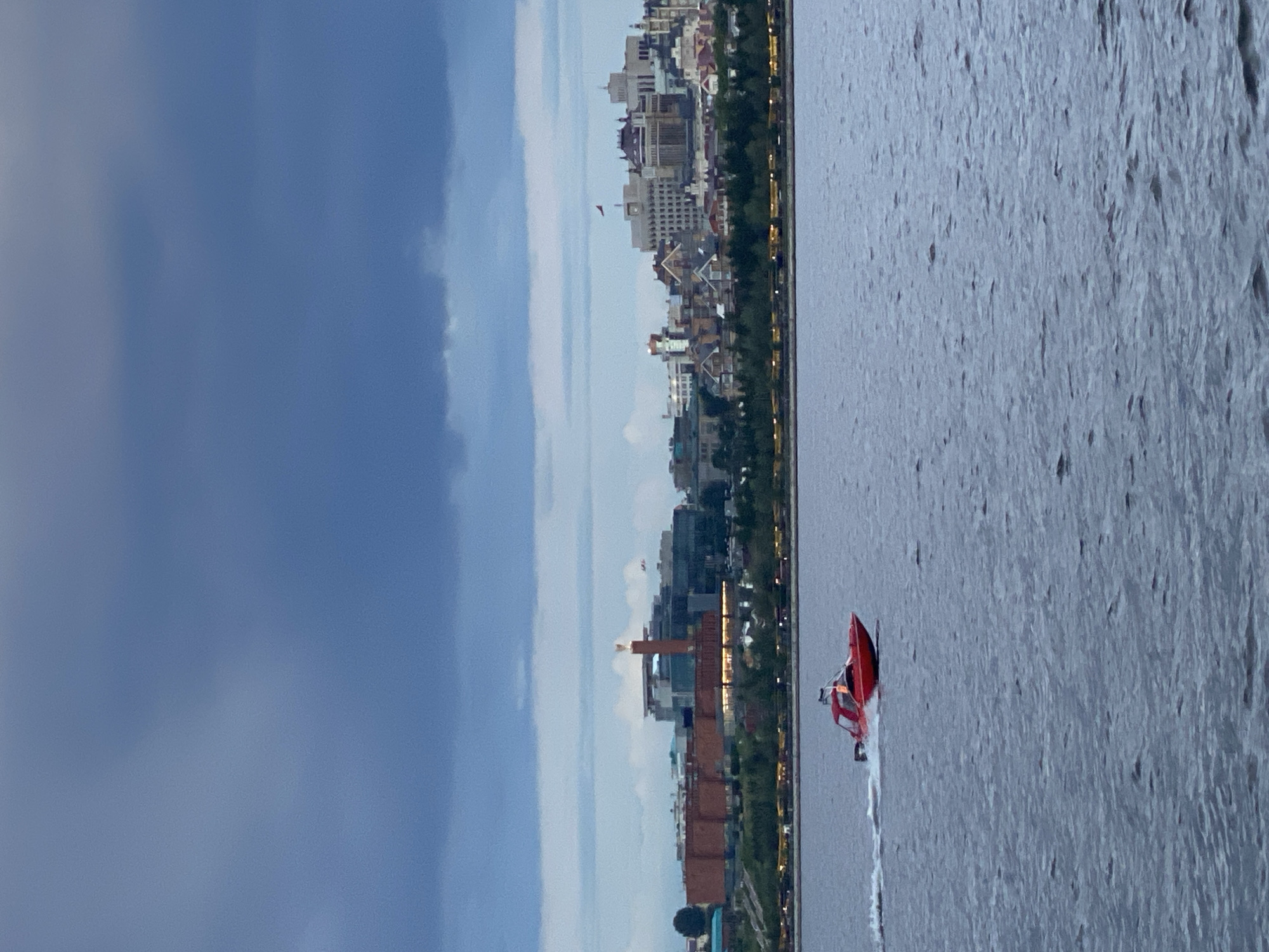 The image, when correctly oriented horizontally, displays a dynamic twilight scene over a body of water, likely a river or large lake, with the city of Kazan, Russia, in the background.
In the foreground, a vibrant red speed boat is actively moving from left to right across the water, generating a clear white wake behind it. The water's surface is textured, reflecting the muted light of the evening.
The midground is dominated by a sprawling city skyline along the embankment. A diverse array of buildings, varying in architectural style and height, lines the waterfront. Many buildings show illuminated windows and facades, and streetlights glow along the water's edge, indicating either dusk or early evening. A strip of green trees separates the buildings from the water.
The background features a dramatic sky, transitioning from a lighter, clearer blue on the right to heavy, dark grey clouds on the left, hinting at changeable weather or the progression of a sunset. The overall mood is serene yet captures a moment of activity. There is no visible text in the image.
