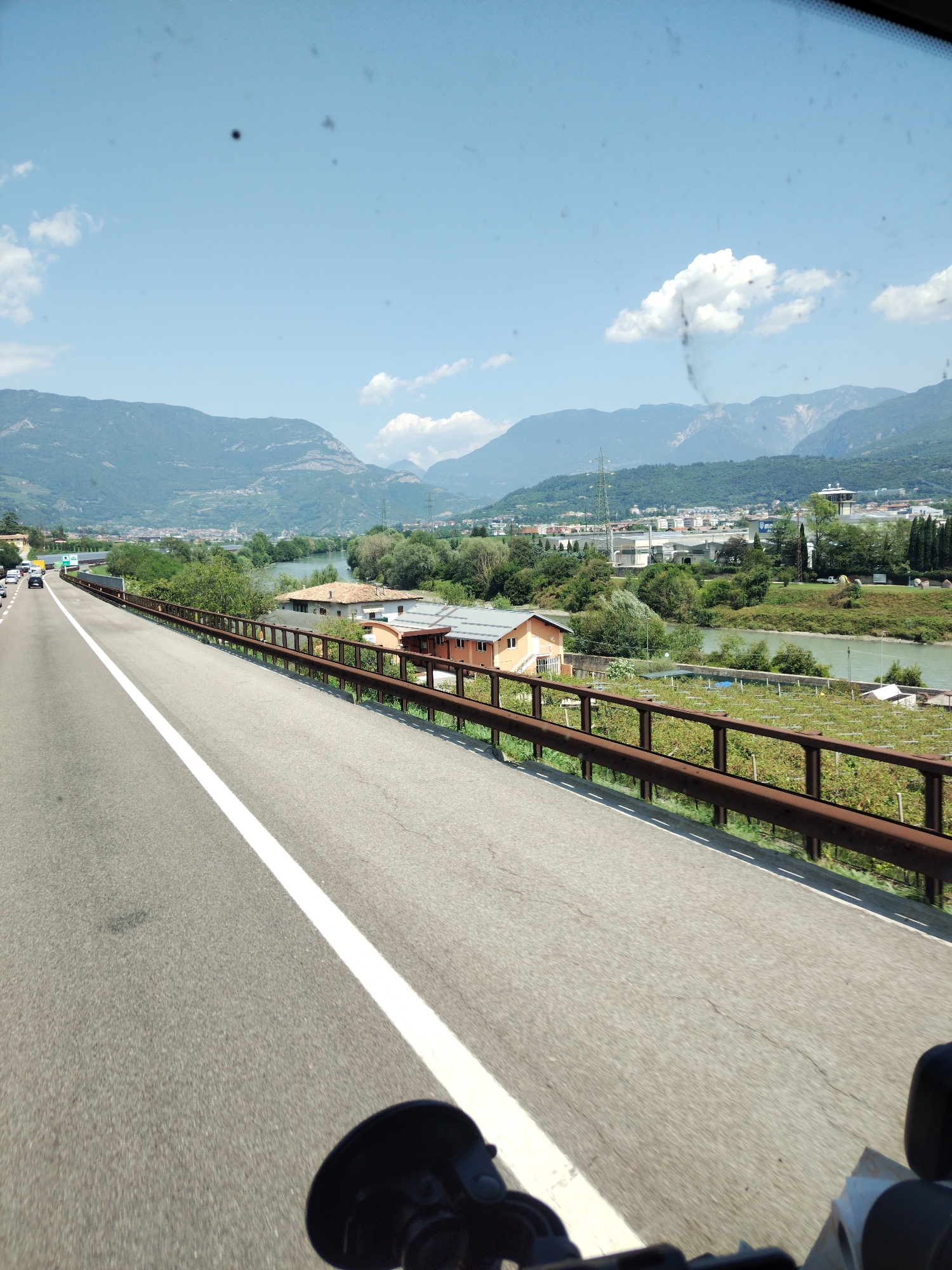 A driver's perspective shot from a vehicle traveling on a highway near Reviano, Italy, shows a wide road with a white lane marker dividing the lanes. The highway is bordered by a rusted metal guardrail on the right. Beyond the guardrail, there are fields with what appear to be vineyards or orchards, a river, and a few buildings with red-tiled roofs. In the background, a town or city is visible nestled in a valley with mountains rising behind it. The sky is blue with scattered white clouds, and the sun is shining. There are no people or specific actions that can be identified in the scene. The image is slightly obscured by dirt or smudges on the windshield.