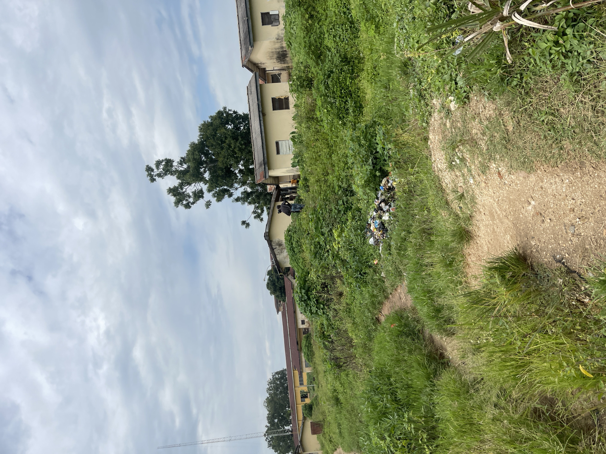 The image depicts a cloudy day in a semi-urban or rural setting, possibly in Jos, Nigeria. In the background, several light-yellow, single-story buildings with corrugated roofs are visible, nestled among lush green vegetation. Some of these buildings appear to be in disrepair, with some windows boarded up or broken. A tall communication tower is present on the left side of the frame, further suggesting a developed, though perhaps not densely populated, area.
In the middle ground, two individuals, dressed in dark clothing, are standing near one of the buildings. Their posture and placement suggest they are observing something or engaged in a casual conversation. In front of them, a pile of assorted trash, including plastic bags and debris, is scattered on the ground amidst the overgrown grass and plants. A dirt path winds through the foreground, bordered by tall, green grass and other vegetation. The overall impression is one of a somewhat neglected environment with signs of habitation and human activity, juxtaposed with natural overgrowth and waste.