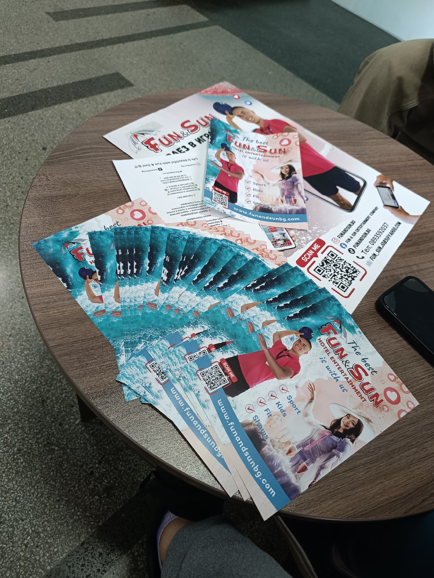 The scene depicts a round, dark wood-grain table upon which numerous promotional flyers for "Fun & Sun," a hotel entertainment company, are arranged. The flyers are designed with colorful graphics including ocean imagery, photos of people, and QR codes. A black smartphone rests on the table to the right. A person wearing tan pants and a gray sweatshirt is partially visible on the right side of the frame. The floor visible in the lower-left corner is tiled in a speckled beige pattern. The flyers include contact information and a website address "www.funandsunbg.com", suggesting a possible location or connection to Bulgaria, Pleven may be a region within Bulgaria.