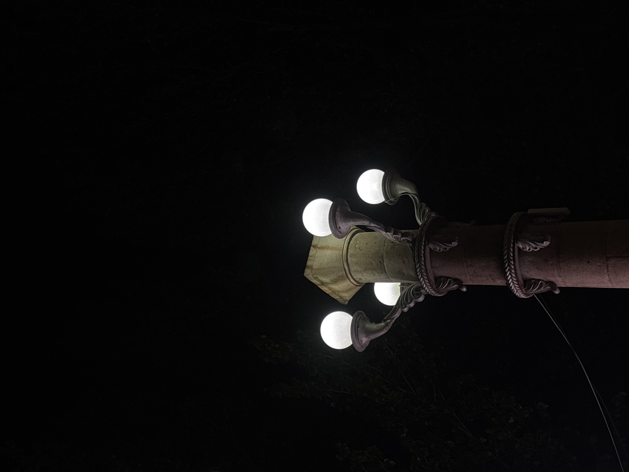 A low-angle shot shows an illuminated, ornate street lamp with five globe lights, positioned against a dark night sky in Gyumri, Armenia. The lamp's support structure is a cylindrical post with decorative rings. Tree branches are barely visible in the lower-right corner. The main focus is on the lamp and its lights. There is no evidence of people or other subjects interacting with the object.