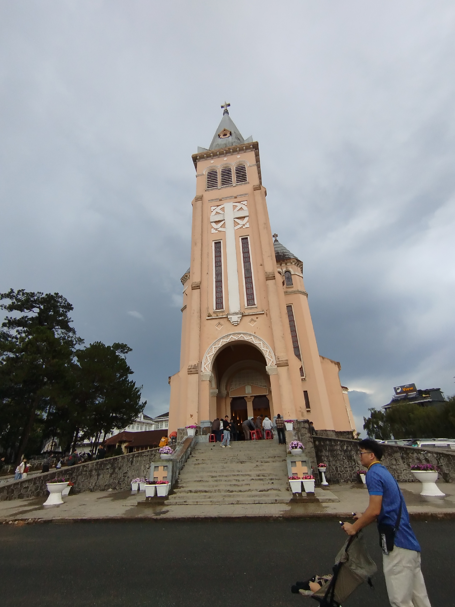 The image shows the Cathedral of Da Lat in Vietnam, a Roman Catholic cathedral. The building has a tall, peach-colored tower with a white cross. A flight of stairs leads up to the entrance, and there are white flower pots with purple flowers lining the steps. Several people are visible on the steps and inside the entrance. A man wearing a blue shirt and light-colored pants is pushing a stroller on the street in the foreground. The sky is overcast with dark clouds. A building with the word "View" is visible in the background. Trees and other buildings surround the cathedral.