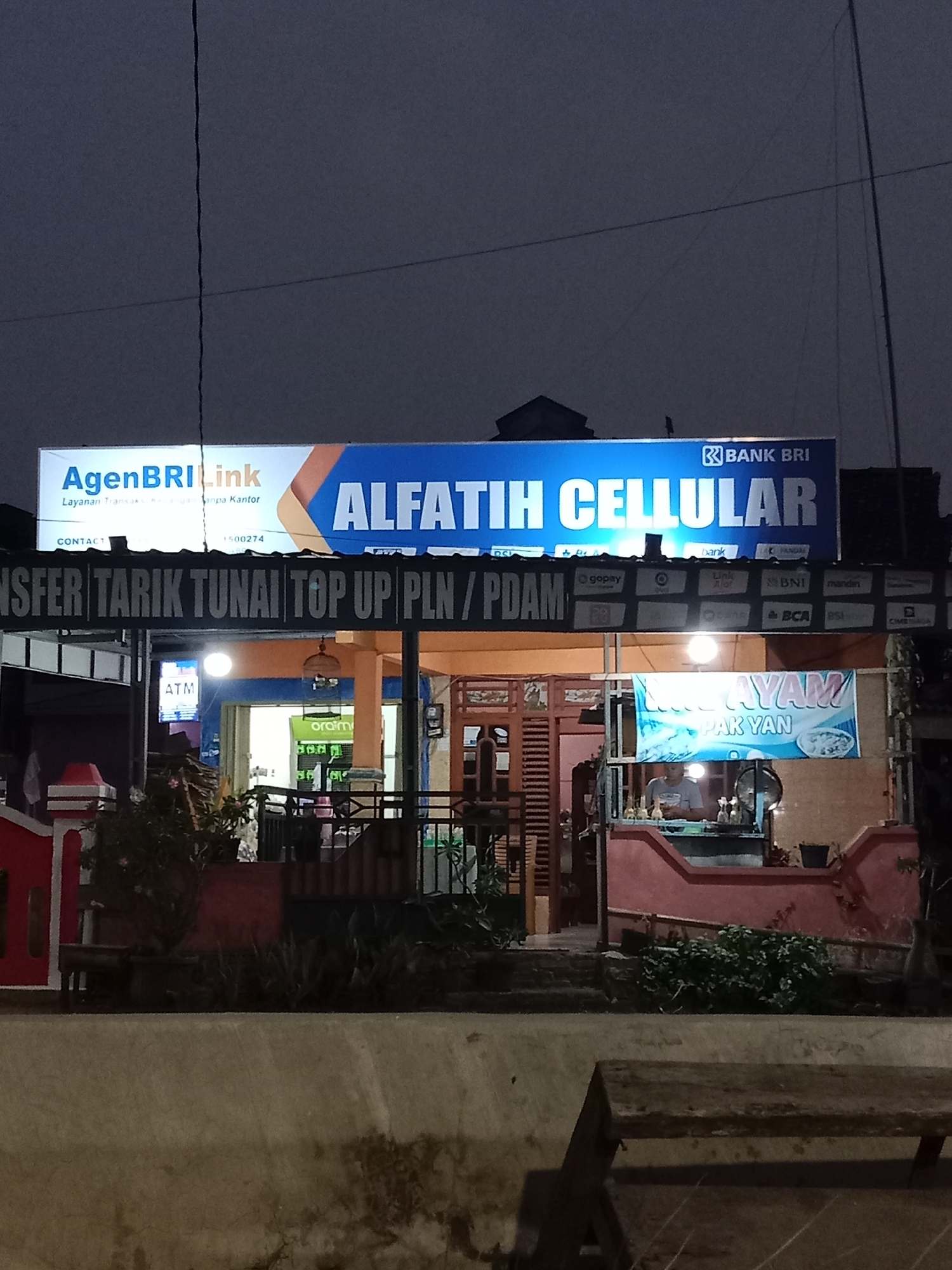 This is a nighttime, eye-level, medium shot of a storefront in Langensari, Indonesia. The storefront has a large illuminated sign that reads "AGENBRI LINK" and "ALFATIH CELLULAR". Below this, a dark sign lists services such as "TRANSFER TARIK TUNAI TOP UP PLN / PDAM". The storefront appears to be a small business with a person visible inside behind a counter, preparing food. A banner advertising "AYAM" (chicken) is also displayed. Various logos of payment providers like "Gopay", "BNI", and "BCA" are visible on the storefront. The environment is dimly lit, suggesting it is evening or nighttime.