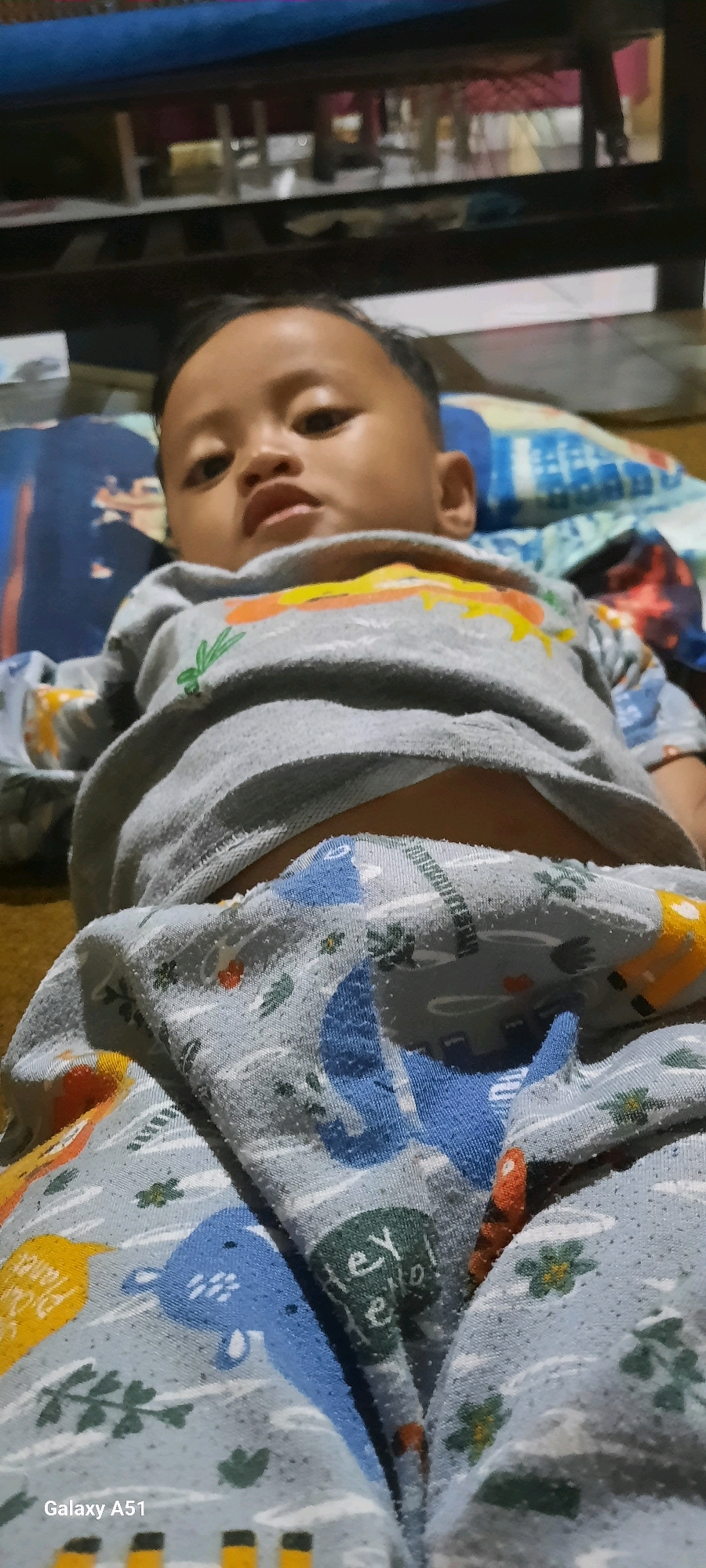The image shows a close-up, low-angle shot of a young child lying down. The child is wearing a grey long-sleeved shirt with cartoonish orange and yellow designs, and light blue pajamas with a pattern of cartoon animals, including what appears to be elephants and perhaps other creatures. The child's face is visible, looking towards the camera with a neutral expression. The background consists of bedding and what looks like furniture, suggesting an indoor setting, likely a home in Jatitengah, Indonesia. The lighting is somewhat dim and appears to be artificial.