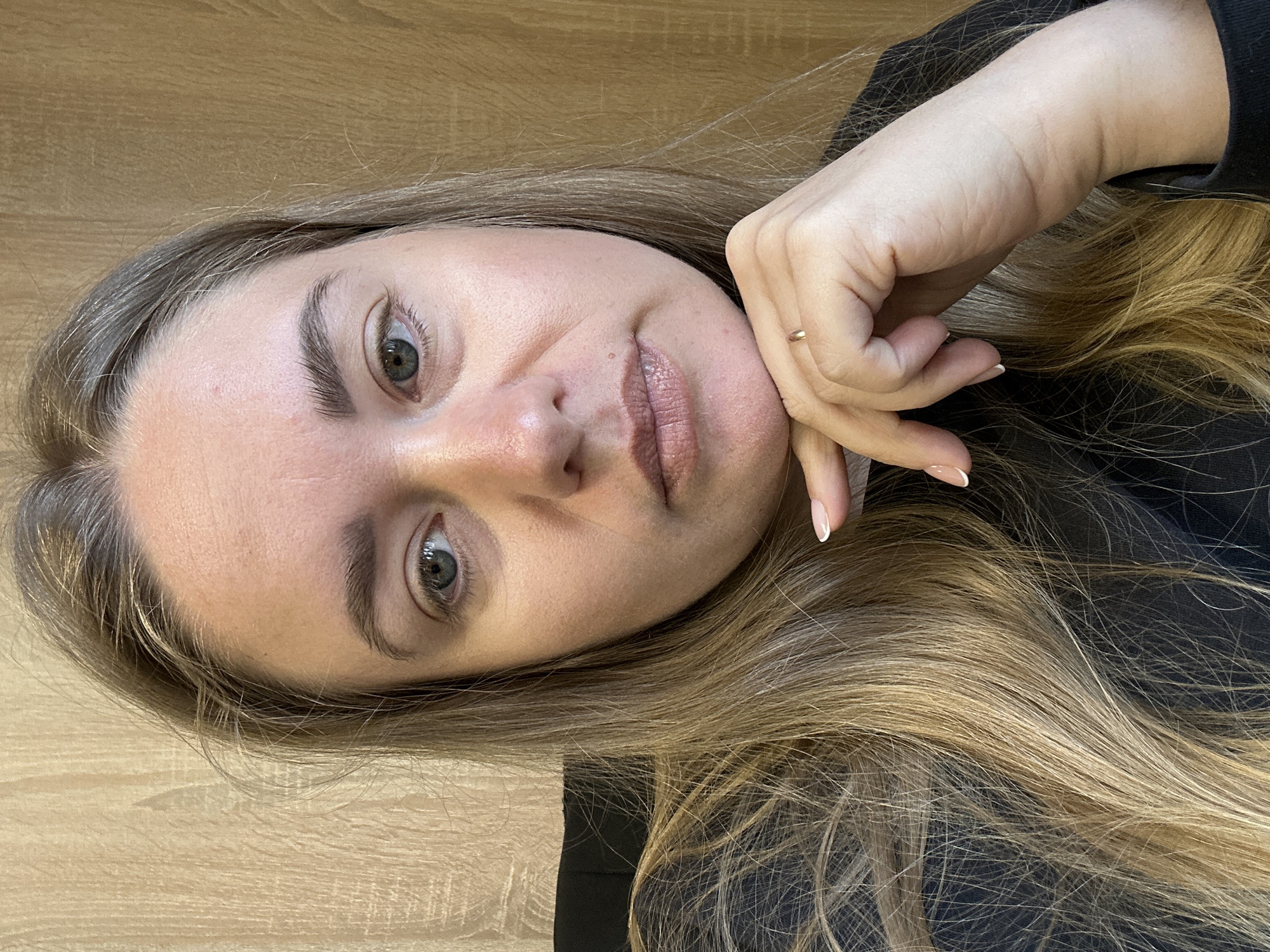 A woman with long, blonde hair and grey eyes is posing for a close-up portrait. Her skin tone is fair. She is wearing a black top. Her left hand is near her face, with her index finger extended along her cheek. She is wearing a gold ring. The background appears to be a wooden surface. There is no indication of the location being Ural'skoe, Russia.