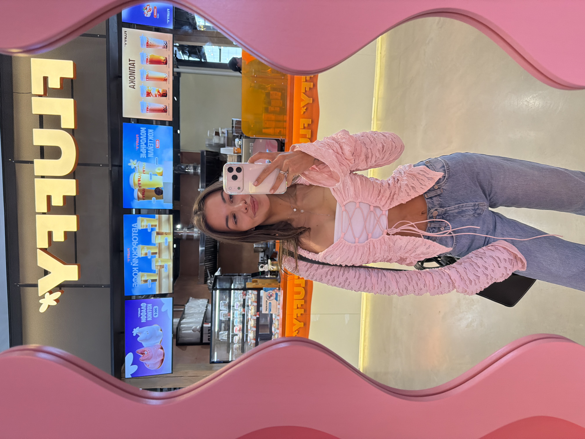 A young woman is captured taking a mirror selfie inside what appears to be a modern cafe or beverage shop in Krasnoyarsk, Russia. She is reflected in a distinctive mirror with a wavy, pink frame. The woman, likely in her early twenties, has long dark hair and is smiling subtly at her phone, which she holds in her right hand. She is dressed in a light pink, textured long-sleeved top with a white lace-up detail over a bralette, paired with light blue jeans. A delicate necklace and a ring on her left ring finger add to her casual yet stylish ensemble.
The background, seen through the mirror's reflection, features digital menu screens displaying vibrant images of various beverages, including bubble teas, milkshakes, fruit teas, and different coffee drinks. A prominent yellow sign, partially reflected, spells out "FREEEYA" (or a similar stylized brand name) and includes a small butterfly or flower logo.
Visible text on the menu screens is primarily in Russian Cyrillic:
* "АКЦИОНПАТ" (AKTSIONPAT), meaning "Promotions" or "Special Offers."
* "МОЛОЧНЫЙ КОКТЕЙЛИ" (MOLOCHNY KOKTEYLI), translating to "Milkshakes."
* "ФРУКТОВЫЙ ЧАЙ" (FRUKTOVY CHAY), meaning "Fruit Tea."
* "ХОЛОДНЫЙ КОФЕ" (KHOLODNY KOFE), for "Cold Coffee."
* "КОФЕЙНЫЕ НАПИТКИ" (KOFEINY NAPITKI), meaning "Coffee Drinks."
* Other details include "300 ₽" (300 Rubles) as a price and "С МЯКОТЬЮ" (S MYAKOTYU), meaning "with pulp."
* A "COFFEE LIFE" logo is also seen on one of the screens.
The setting suggests a brightly lit, contemporary establishment, indicating either daytime or evening hours. The central activity is the woman taking a self-portrait within this inviting environment.