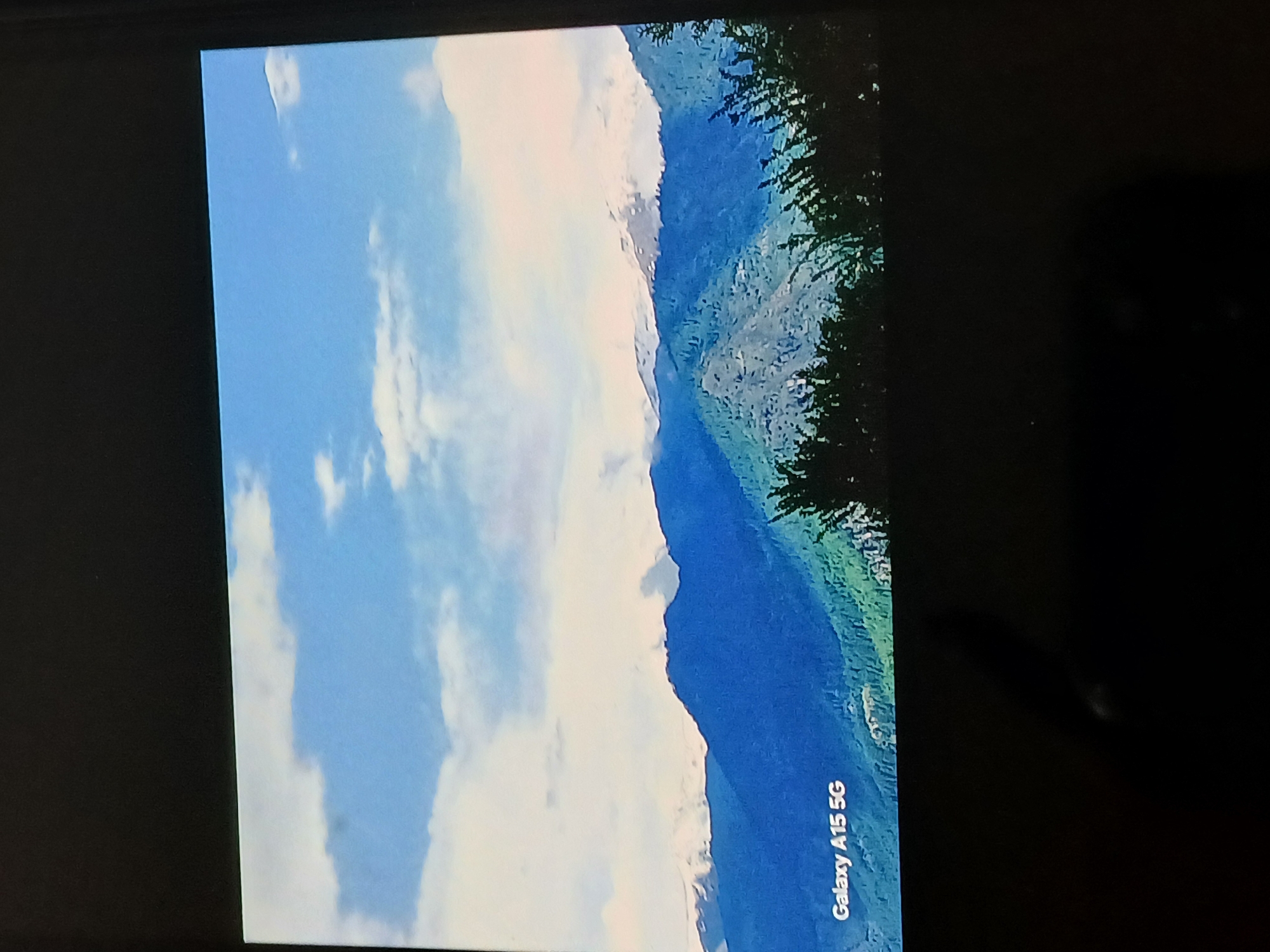 The image shows a mountain landscape with a blue sky and clouds, displayed on a phone screen, possibly a Galaxy A15 5G. The lower right corner of the screen is visible and labeled “Galaxy A15 5G”. The surroundings are dark, focusing attention on the illuminated screen. The image's location is unspecified, and there is no indication of it being in Bengaluru, India.
