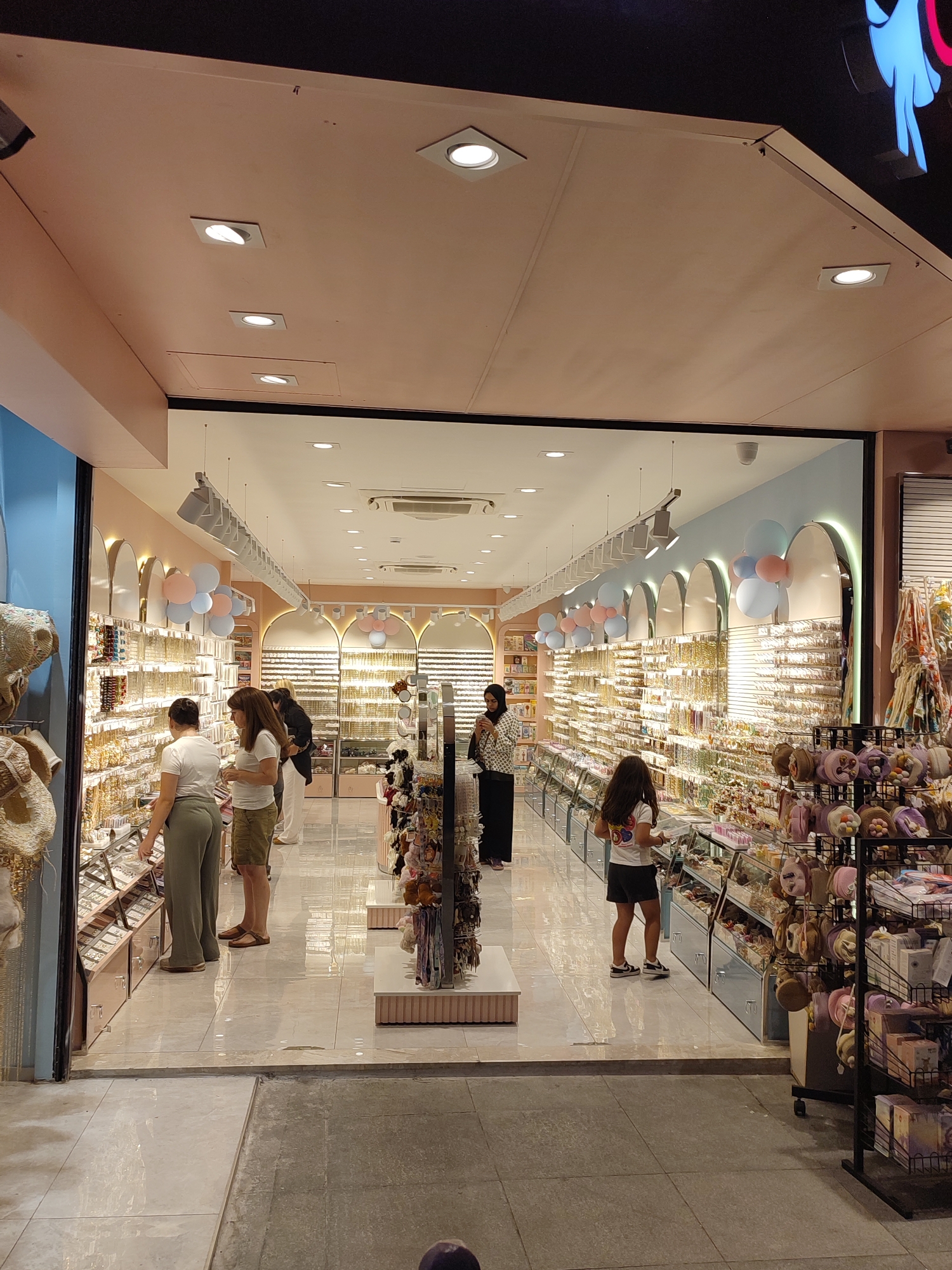 The image shows the interior of a brightly lit retail store, likely a boutique specializing in accessories. The store features extensive display shelves filled with various items such as jewelry, hair accessories, and possibly sunglasses. In the foreground, two women are browsing. One woman, wearing a white t-shirt and olive green pants, is examining items on a display shelf. The second woman, in a white t-shirt and khaki shorts, is standing next to her, also looking at the merchandise. Further into the store, a young girl in a white t-shirt and black shorts is looking at items on display. Behind her, a woman wearing a black jacket and white pants stands near a display. The store's interior design includes pastel-colored walls, arch-shaped display units, and decorative balloons in pink and light blue. The flooring is a reflective tile. The overall impression is of a modern, well-organized retail space. The mention of Kadıköy, Turkey, suggests this is a specific location for this store.