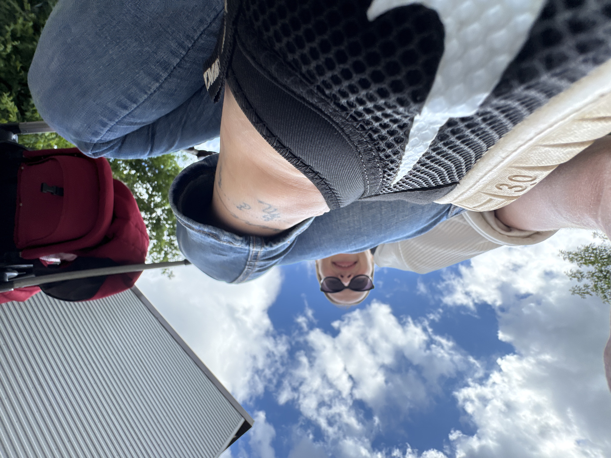 The image shows a low-angle, close-up shot of a person's legs and feet, with a clear blue sky and white clouds visible in the background. The person is wearing blue denim jeans and black mesh sneakers with white accents. A red bag is visible in the upper portion of the image, along with part of a ribbed, light-colored surface that appears to be an awning or roof. A tattoo is visible on the person's ankle. In the distance, trees and foliage can be seen. The perspective suggests the photo was taken by someone lying down or sitting with their legs raised towards the camera. The image does not contain enough information to determine a specific location in Chaikovsky, Russia.