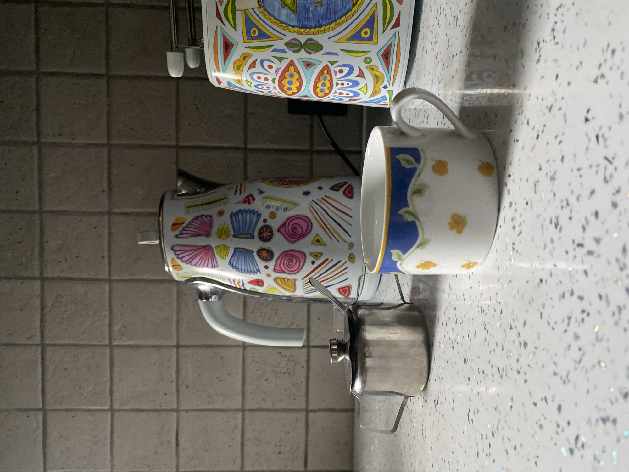 A close-up, eye-level shot captures a kitchen counter scene, focusing on a decorated electric kettle, a small stainless steel sugar pot with a spoon, and a ceramic mug filled with liquid. The kettle, positioned centrally, is adorned with a vibrant, abstract pattern of colorful shells, lines, and swirls. To its left, the sugar pot has a lid and two small handles, with a spoon resting inside. In the foreground, the mug is white with a blue rim and a delicate pattern of green vines and small yellow flowers. The background features beige tiled walls. The countertop is speckled white with reflective particles. A toaster with a similar colorful, geometric design is partially visible on the right. A black electrical cord extends from the toaster towards the back. The lighting suggests an indoor environment, likely a kitchen, casting shadows on the countertop. There is no indication of any specific location in Wrocław, Poland.