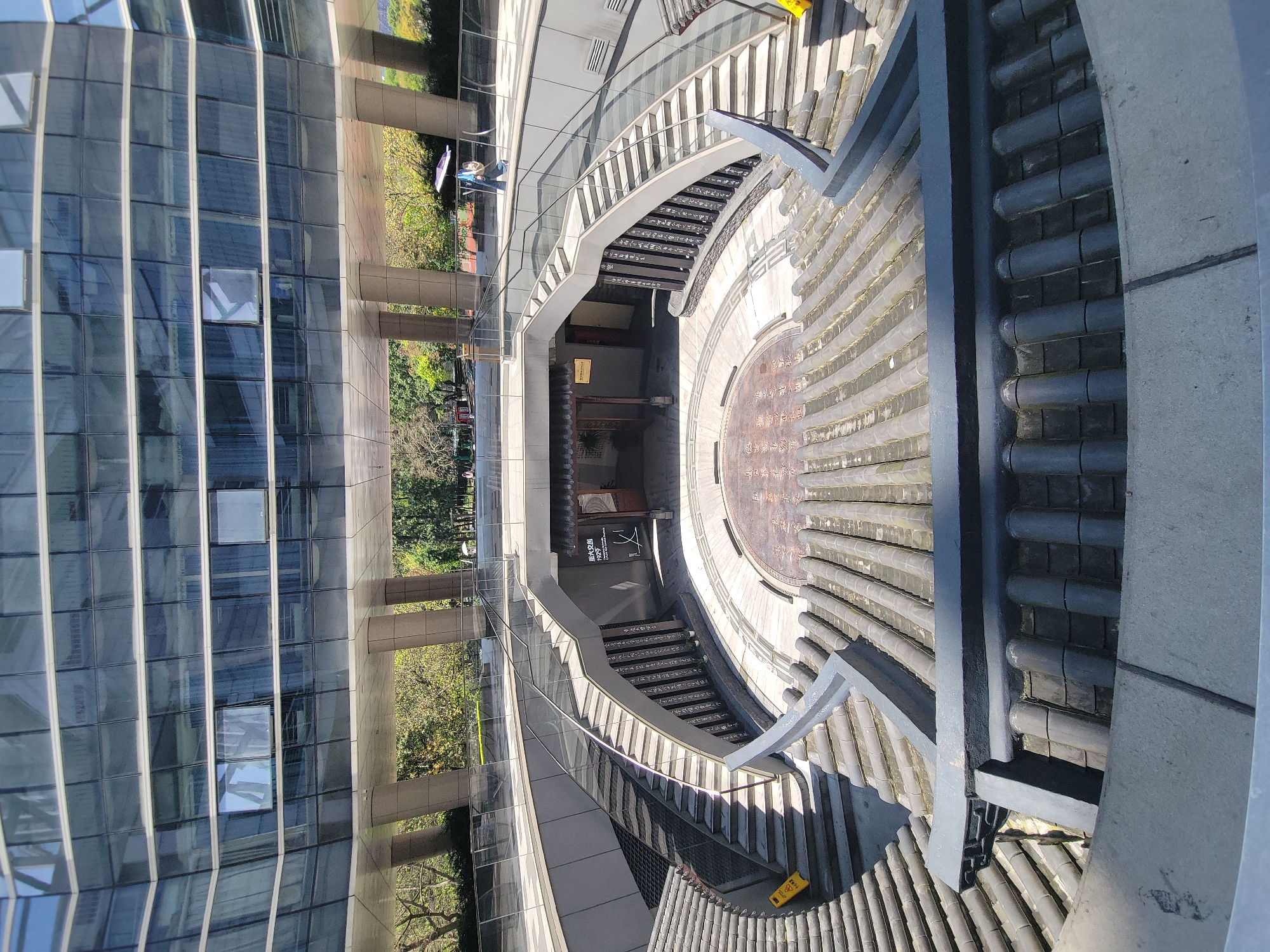 An overhead, high-angle view captures a blend of modern and traditional architecture, likely in Shapingba District, China. To the left, a tall, multi-story building features a contemporary glass facade with numerous square windows, reflecting the surroundings. Several people are visible in the distance, walking outdoors near the base of this building; one person holds an umbrella, possibly for sun protection.
To the right, contrasting sharply with the modern structure, is a circular sunken courtyard or plaza designed in a traditional Chinese style. Its perimeter is lined with dark grey, curved roof tiles, resembling those found on ancient temples or official buildings. A series of stone or concrete stairs, equipped with modern glass railings, descends into this courtyard, revealing intricate architectural details and further tiled roofs at lower levels. A prominent feature in the center of the circular courtyard is a large, round decorative element or plaque covered with Chinese characters, which appear to be part of a historical or commemorative inscription. Phrases like "...人民共和国..." (People's Republic of...) are partially visible on this plaque.
At the lowest level of the sunken area, traditional wooden pillars and beams frame an entrance or structure, with a sign bearing Chinese characters "曾家岩 1955" (Zēngjiāyán 1955) visible. A yellow warning sign is placed on a section of the stairs on the right. Lush green foliage and trees are visible beyond the structures, suggesting an outdoor, possibly park-like, setting. The scene is illuminated by bright daylight, casting distinct shadows and indicating clear, sunny weather. The overall impression is one of a cultural or public space that harmoniously integrates historical aesthetics with modern design.