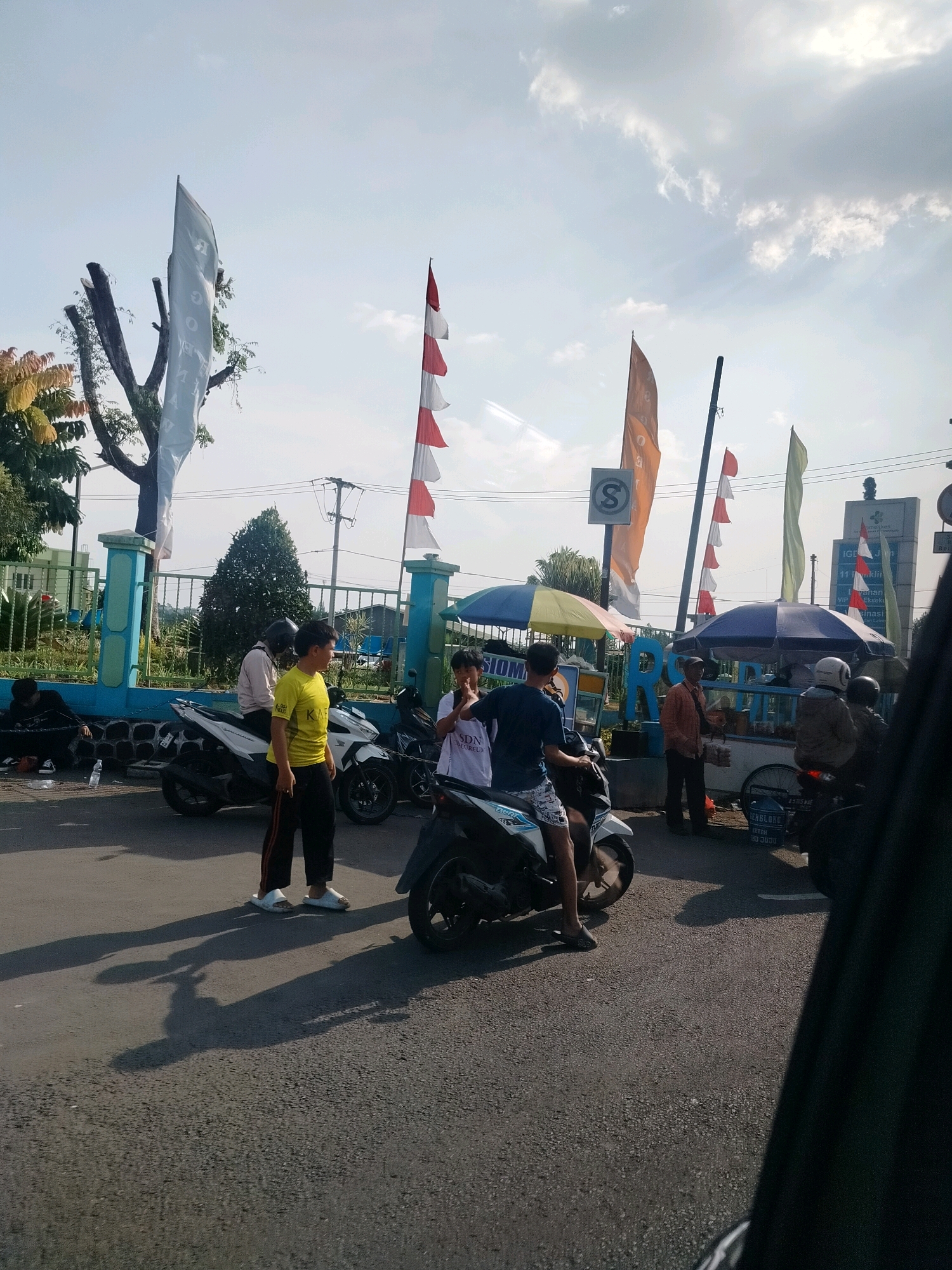 The image depicts an outdoor scene in Cisarua, Indonesia, likely a public area with a road and pedestrian traffic. Several people are present, some on or near motorcycles, suggesting a transportation hub or gathering point. A food vendor with a cart and umbrella is visible, indicating commercial activity. Decorative flags are flying, some with red and white stripes, possibly nationalistic symbols or event markers. The environment appears to be a sunny day with some clouds in the sky. The overall impression is a snapshot of everyday life in a busy urban or semi-urban setting in Indonesia.