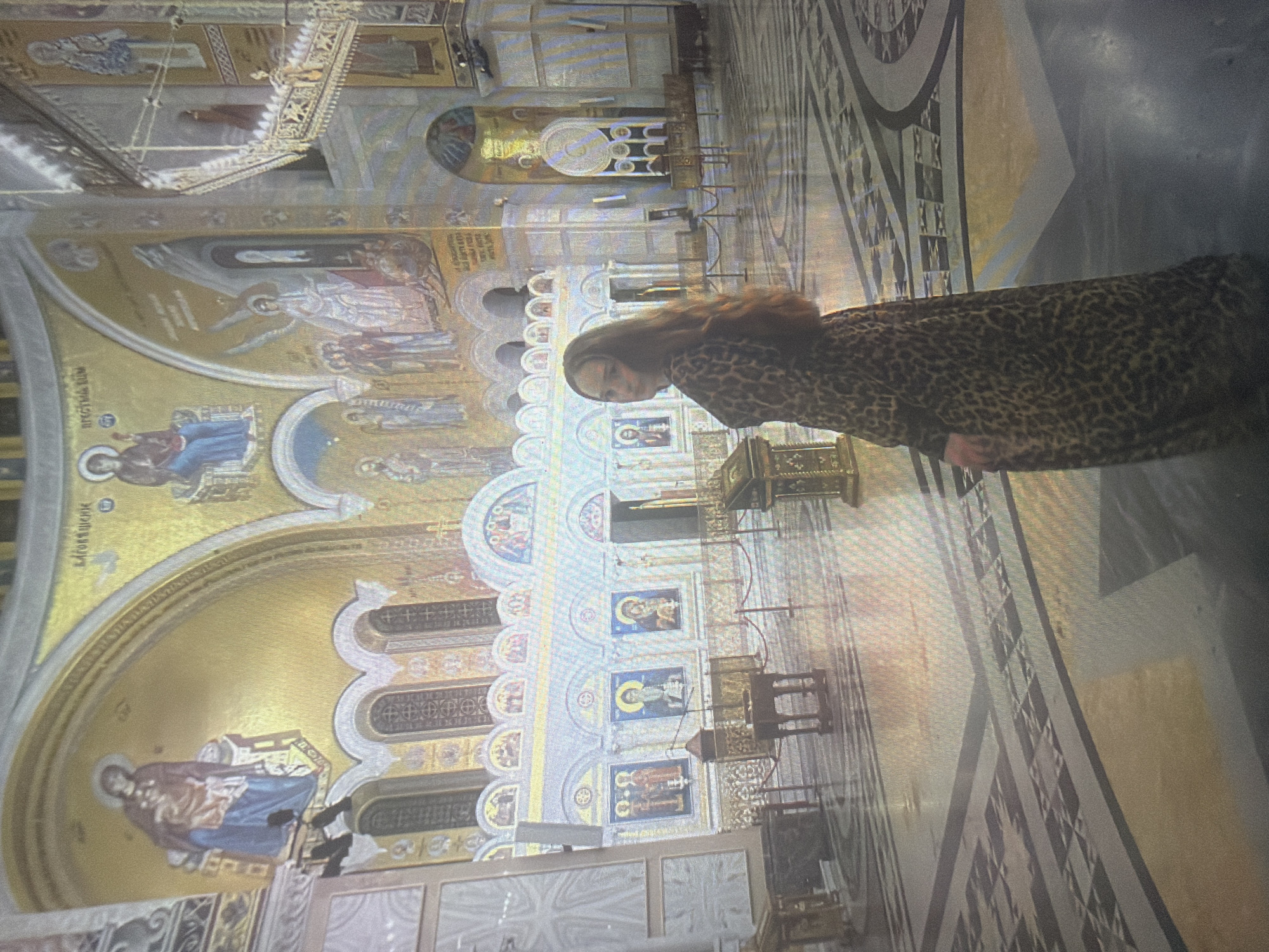 The image depicts the interior of a large, ornate Orthodox Christian cathedral in Shumen, Bulgaria, characterized by extensive gold mosaics and polished stone surfaces. A single female individual is centrally positioned, facing away from the viewer but turning her head and upper torso to look over her right shoulder towards the camera. She has long, wavy brown hair and wears a full-length, long-sleeved dress with a brown and black leopard print pattern.
The cathedral interior features towering walls and arches adorned with intricate gold mosaics. On the upper left, a mosaic depicts a female figure in red and blue robes, with Cyrillic text reading "БЛАГОВЕЩЕНИЕ" (Annunciation) above. In the upper middle section, another mosaic portrays a seated female figure, likely the Virgin Mary, with Cyrillic text "ПРЕСВЕТА БОГОРОДИЦА" (Most Holy Mother of God) above. Adjacent to this, a large mosaic of an angel is visible. The lower walls are decorated with arched sections containing rectangular mosaic icons of saints.
To the right, an elaborate multi-tiered structure, possibly a pulpit or part of an altar screen, features ornate gold metalwork and additional mosaic details. The floor is constructed from large, highly polished stone tiles arranged in a complex geometric pattern of light grey, dark grey/black, and gold/light brown segments, reflecting the ambient light. Several wooden pedestals or offering tables and a dark, ornate wooden lectern are present in the mid-ground. Areas are delineated by red velvet ropes suspended between thin gold stanchions. The entire space is brightly illuminated, casting a warm, golden ambiance.