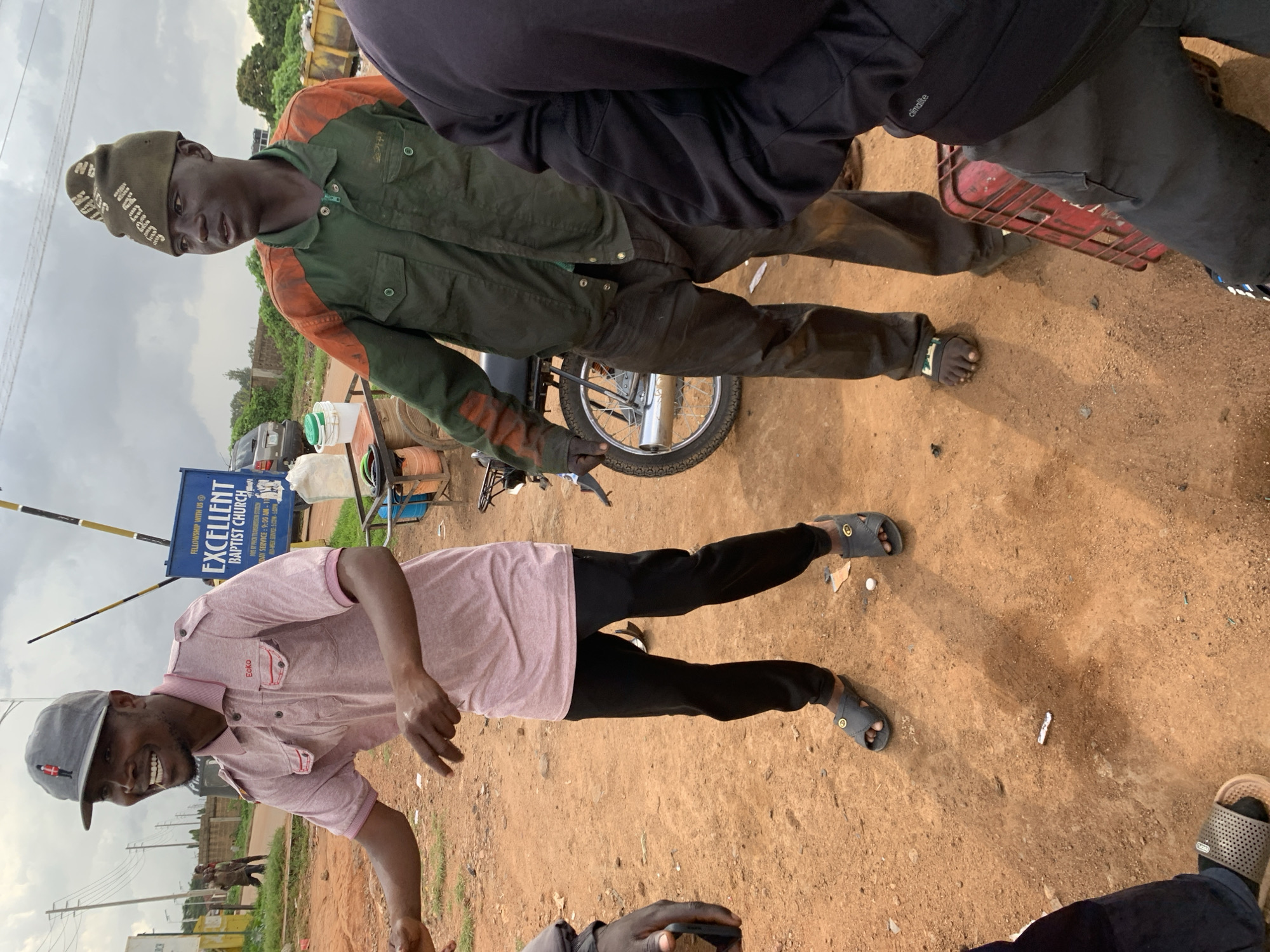 In Barakin Lerawi, Nigeria, two men are standing outdoors on a dirt ground. The man on the left is wearing a pink polo shirt and black pants, with a grey baseball cap. He is smiling and gesturing with his hands. The man on the right is wearing a green and orange jacket and dark pants, with a dark green beanie. He is looking to the side. Behind them, a blue sign reads "EXCELLENT BAPTIST CHURCH." A motorcycle is visible to the right of the man in the green and orange jacket. The ground is covered in reddish-brown dirt and there are some small white objects scattered around. The sky is overcast.