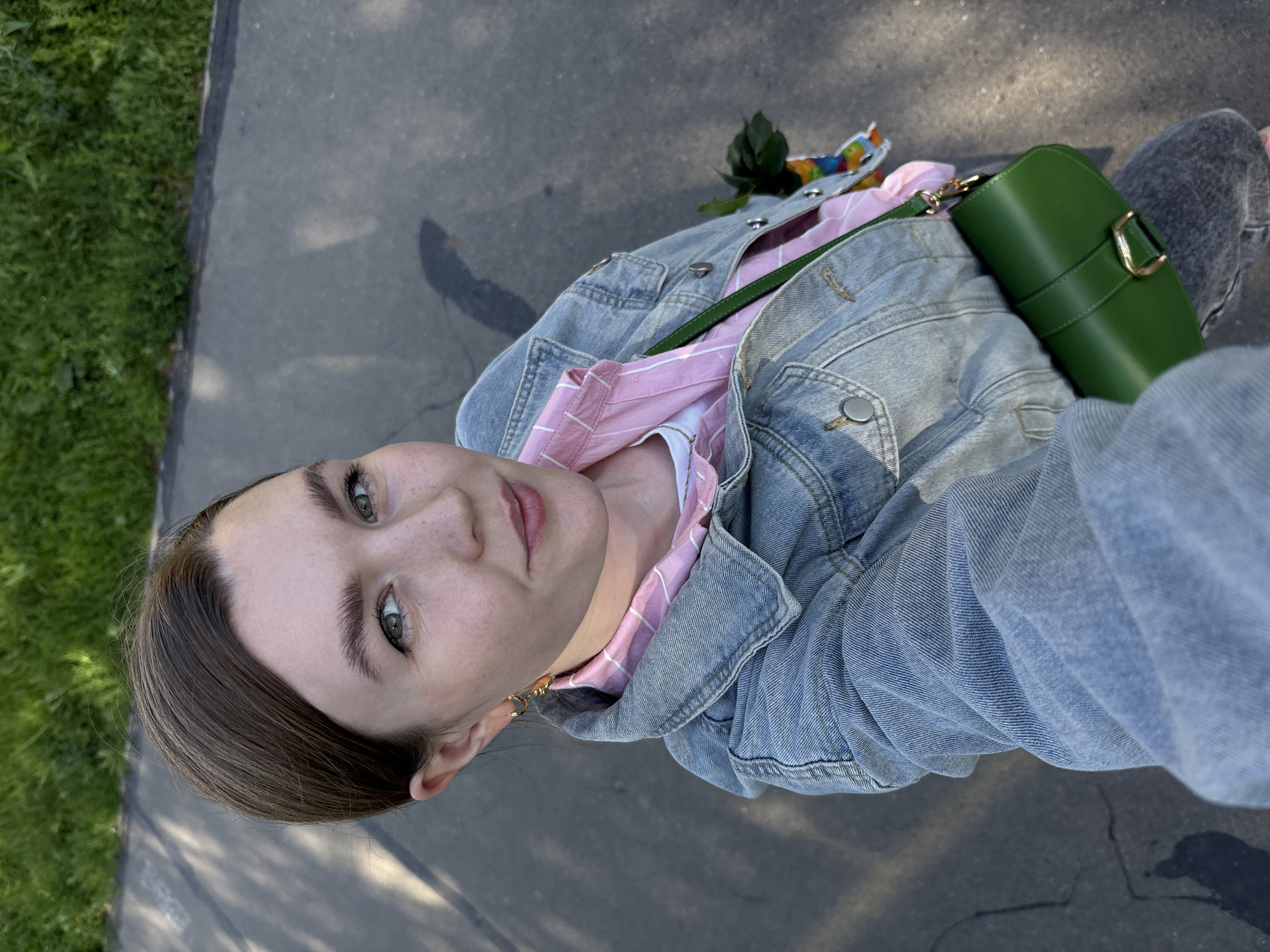 A high-angle, eye-level, medium outdoor selfie shows a young woman with brown hair pulled back, fair skin, and green eyes. She is wearing a light blue denim jacket over a pink and white striped shirt, and a green purse with a gold buckle is slung across her body. She is holding a small bunch of dark green leaves with a colorful lanyard in her left hand. The background shows a paved pathway with shadows and a strip of green grass. The woman appears to be walking on a sunny day in a park or along a street. The image is likely taken in Izhevsk, Russia, given the context.