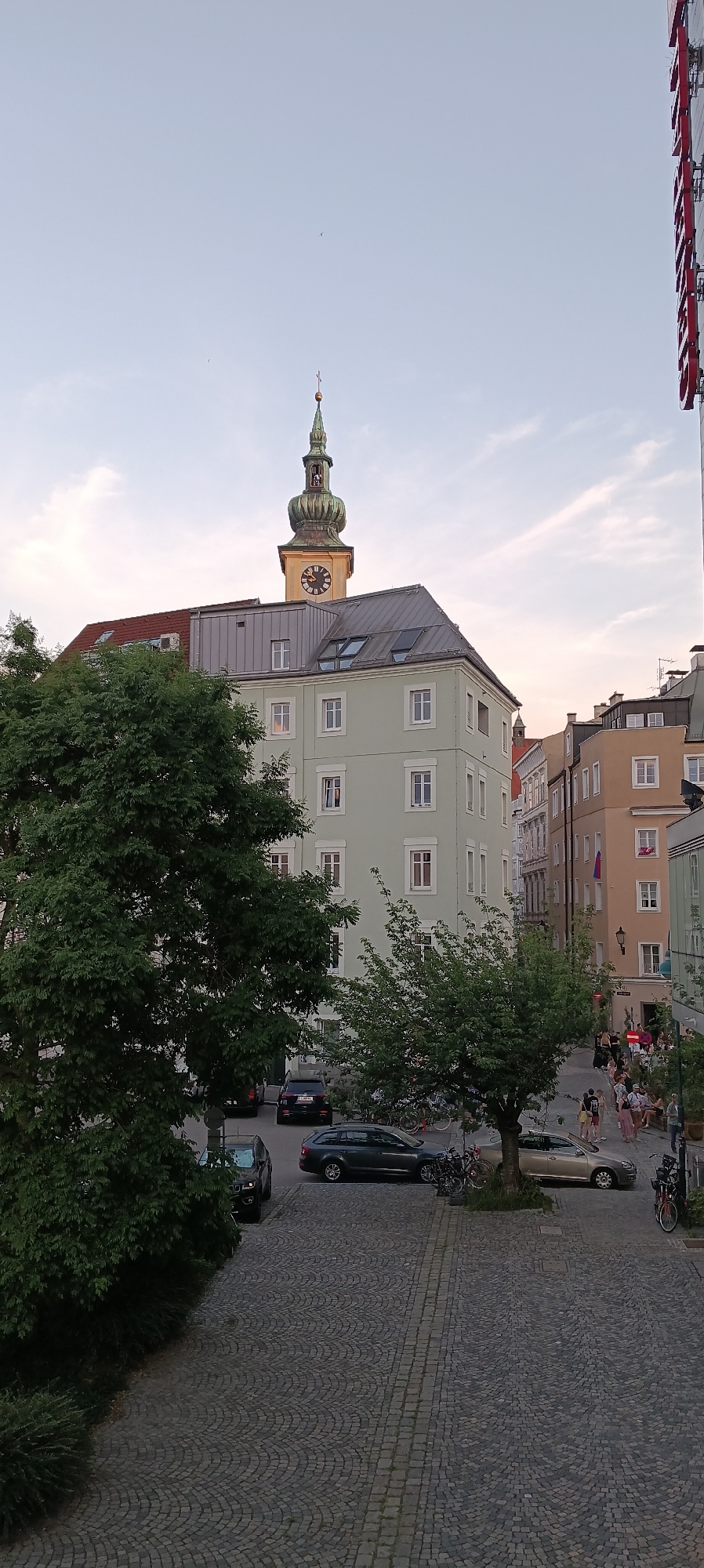 This is a vertical, medium-angle, exterior, full-color photograph captured in Linz, Austria, looking towards a street scene with buildings and a church tower. The time of day appears to be late afternoon or early evening, with soft, diffused light and a hazy, pale blue sky with wispy clouds.
In the foreground, a cobblestone-paved area with a curving pattern leads towards a street. A large, dark green tree with dense foliage occupies the left side of the frame, partially obscuring the view of the buildings behind it.
The midground features several buildings. A light green building with multiple windows and a grey, sloped roof dominates the center. To its right, a series of taller, older buildings with varying facades and windows line the street. One of these buildings displays a small Austrian flag. A prominent church tower with a green, multi-tiered spire and a clock face stands tall above the buildings in the background.
In the street, there are several parked cars, including a dark colored SUV and a silver sedan. A few bicycles are also visible. A group of people, appearing to be casually strolling or socializing, are gathered on the sidewalk in the distance, near the buildings. The overall impression is of a typical European city street.