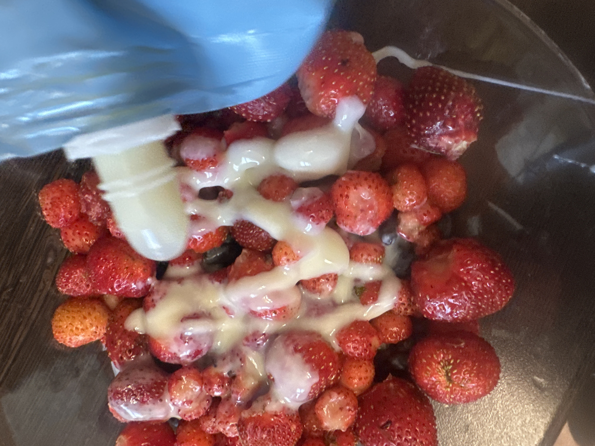 A close-up, overhead shot shows a bowl of strawberries being drizzled with a creamy white liquid from a blue dispenser. The strawberries vary in size and ripeness, with some appearing bright red and plump, while others are smaller and slightly more orange. The liquid coats the berries, creating a glossy sheen. The background is a dark, textured surface, possibly a wooden table. The overall scene suggests a preparation of a dessert or a simple snack. This image does not contain any identifiable landmarks or details to ascertain its location in Novoaltaysk, Russia.