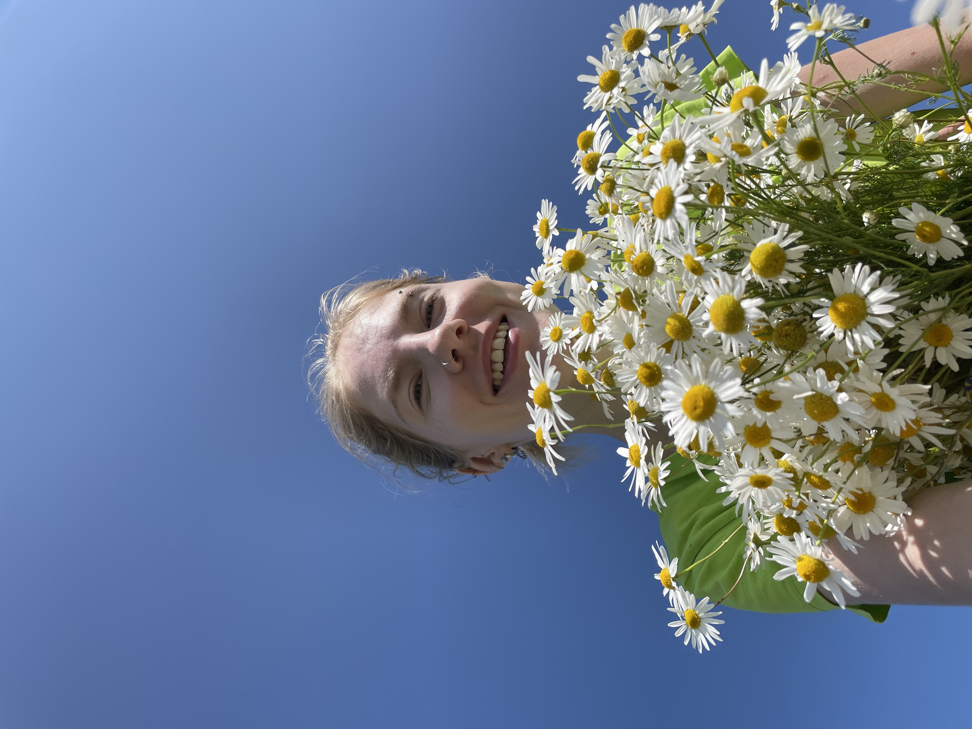 In the city of Сям-Можга, Russia, a young woman with blonde hair, wearing a bright green shirt, smiles while holding a large bouquet of white daisies with yellow centers. She has a nose ring and several earrings. The flowers are in the foreground, obscuring much of her body. The background is a clear, bright blue sky, indicating an outdoor setting on a sunny day. The image is captured from a low angle, looking upwards at the woman and the sky.
