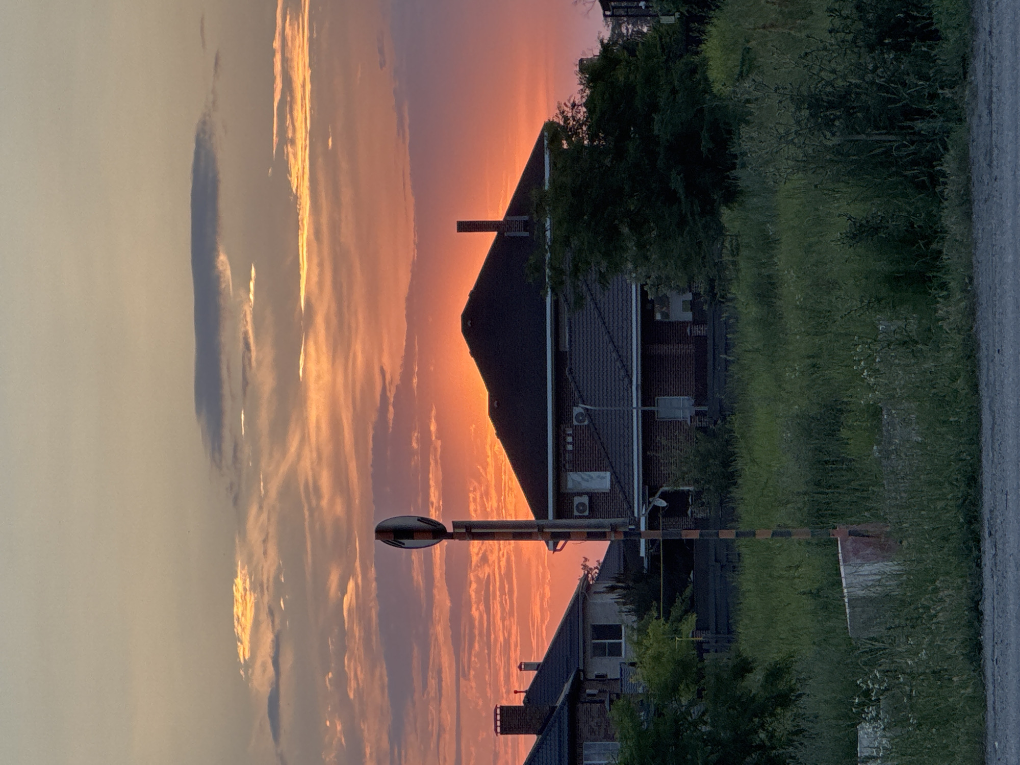 The image captures a suburban scene at dusk, with a vibrant sunset dominating the sky. The sky is ablaze with hues of orange, pink, and yellow, with wispy clouds scattered across it. In the foreground, a tall pole with a circular, checkered marker stands prominently. Behind this pole, several houses with dark tiled roofs are visible. Some houses are made of brick, while others appear to have lighter colored facades. Trees and dense green vegetation surround the houses, partially obscuring them. The overall impression is one of a quiet residential area bathed in the warm glow of the setting sun. The location is identified as Unknown, Russia.