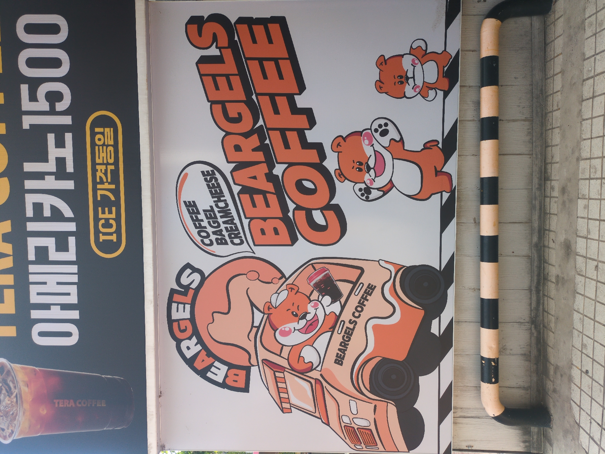 This image depicts a promotional sign for "BEARGELS COFFEE," a coffee shop that also sells bagels and cream cheese. The sign features cartoon bears, with one driving a car labeled "BEARGELS COFFEE" and holding a beverage. Another bear is waving. The sign also advertises "ICE," likely referring to iced coffee, and lists a price of 1500 (Korean Won). A photograph of iced coffee is included. There is a black and cream striped poll next to the sign. The sign is likely located in Seoul, South Korea, based on the Korean language text.