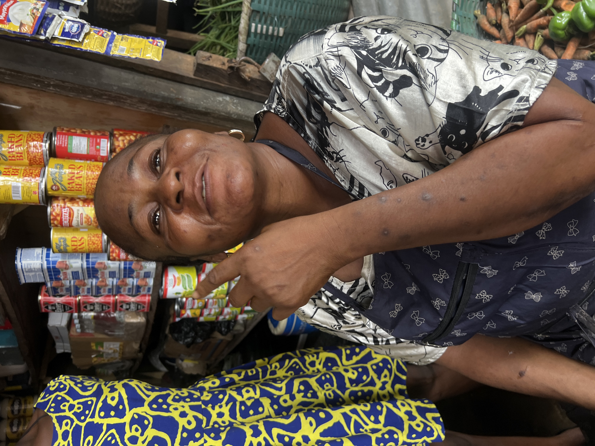 A middle-aged Black woman, with dark skin, smiles at the camera in a marketplace in Nkpor, Nigeria. She is wearing a white shirt with a black cat print and a dark blue apron with a bow pattern. Her left arm is bent, and she is pointing to the right with her index finger. She has on a gold hoop earring in her right ear. Behind her, shelves are stacked with canned goods and other merchandise. To her left, another person, partially visible, wears a blue and yellow patterned dress. In the background, a basket of carrots and green peppers is visible. The overall setting appears to be a small, open-air stall or shop.