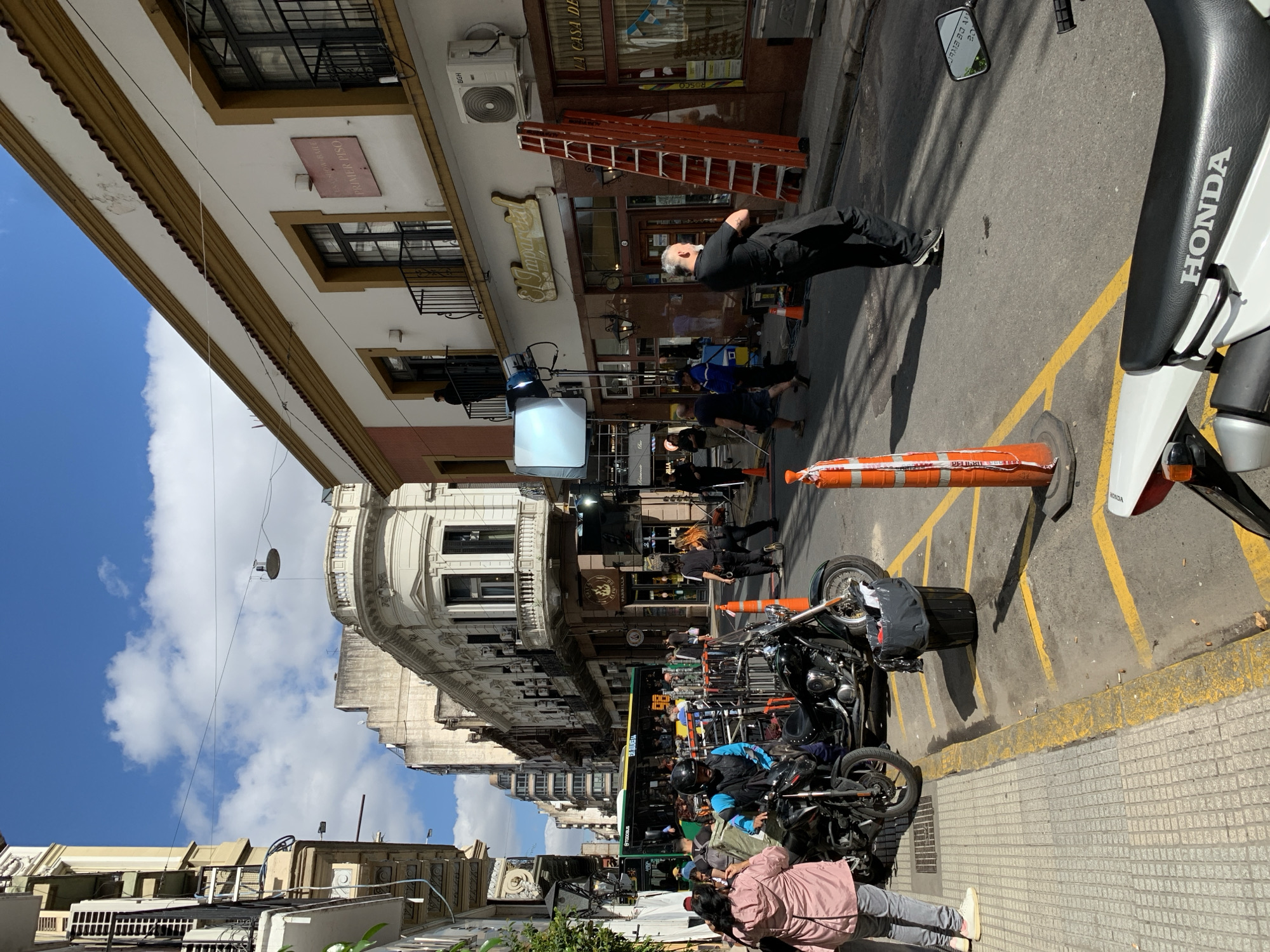 The image captures a street scene, possibly a film set, in Buenos Aires, Argentina. The environment is urban with buildings of classical architecture along the street. A bus is visible amid many motorbikes. An orange ladder is positioned outside one building. There are a number of people in the shot, including one reclining on the pavement outside a building with a sign that says ‘El Imparcial.’ A film crew and lighting equipment, including a large softbox, are evident. A Honda motorbike is in the foreground. The sky is partly cloudy with blue showing.