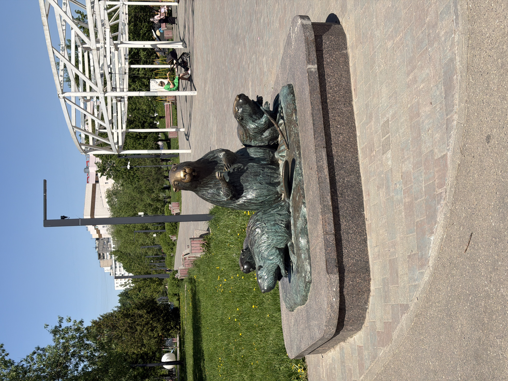 A bronze statue of beavers stands in a park-like area in Moscow, Russia, on a sunny day. The sculpture, depicting a family of beavers, rests on a stone base. A pedestrian walkway of brick and concrete surrounds the statue. In the background are trees, benches, and an open, white metal structure. Lampposts are positioned along the walkway. Buildings are visible in the distance. A person in green is swinging on the white metal structure.