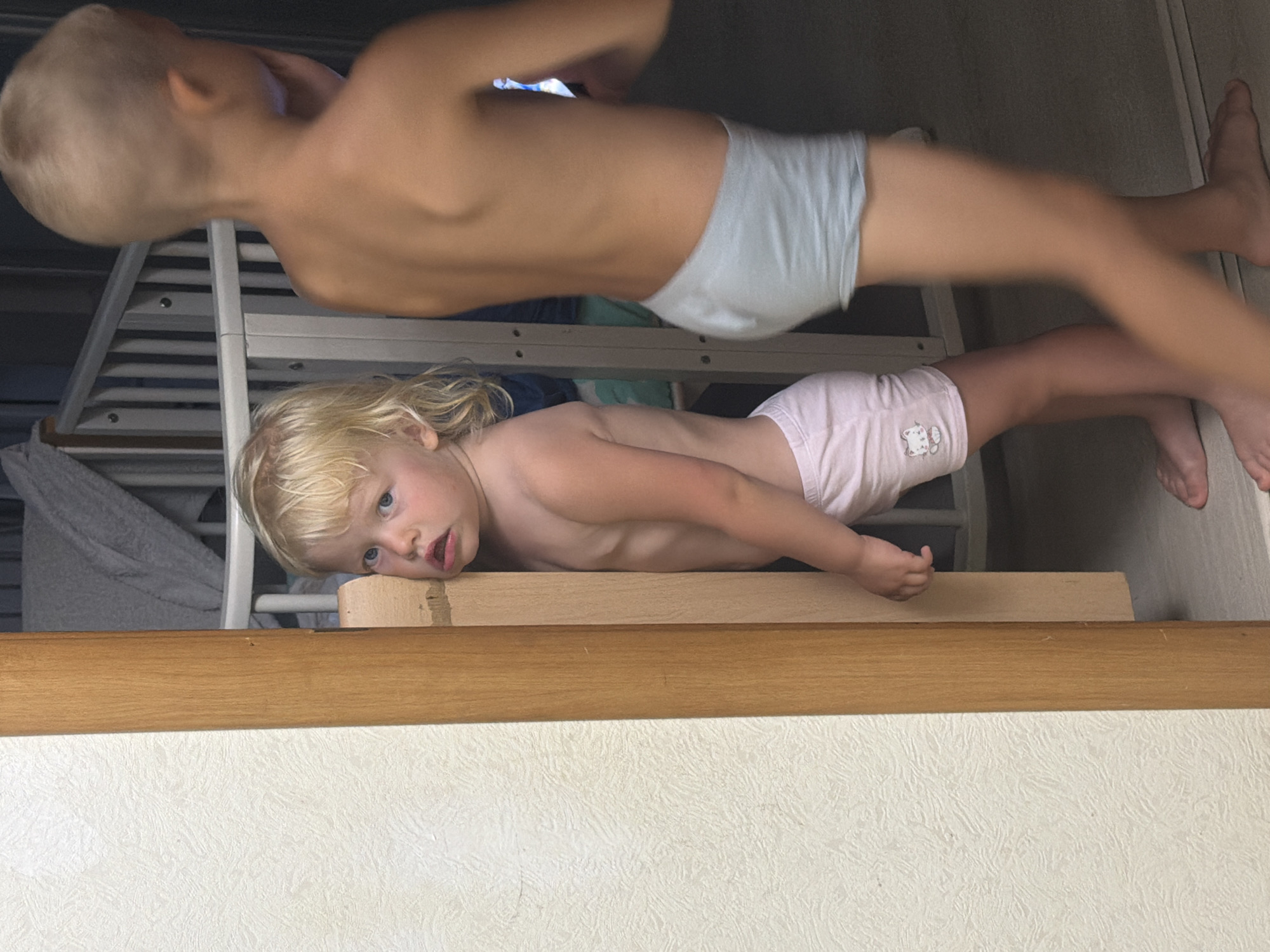 The media file shows two young children, a boy and a girl, in what appears to be a domestic setting. The image is oriented vertically, with the subjects positioned in the foreground and middle ground.
The boy, who is closer to the top of the frame and positioned behind the girl, has blonde hair and is wearing light blue underwear. He is lying down, with his back to the camera, and his body is mostly visible from the waist up. His skin tone is light.
The girl, positioned in the lower half of the frame, has blonde hair and blue eyes. She is wearing pink underwear with a small white cat graphic on it. She is lying on her side, facing the camera, with her head propped up on a wooden surface. Her expression appears curious or possibly a little bewildered.
The background is blurred but suggests an indoor environment with some kind of multi-tiered structure, possibly a bunk bed or crib, and a wall with a textured surface. A wooden plank runs horizontally across the lower part of the image, separating the lower portion of the frame from the upper.
The lighting suggests it could be daytime, possibly indoors with natural light. No specific time of day or weather conditions are discernible. The overall scene has a candid, everyday feel, capturing a moment of rest or play for the children. No text is visible in the image.