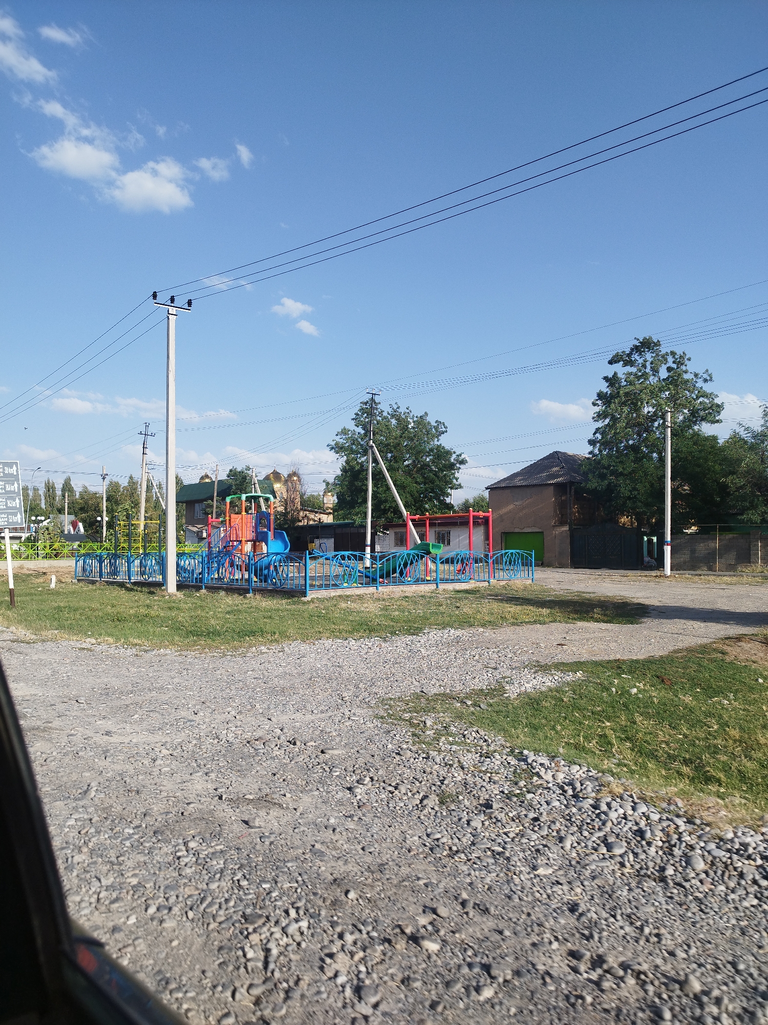 The image captures a street scene in Зертас ауылдық округі, Kazakhstan, on a clear day with a blue sky and scattered white clouds. In the foreground, a gravel road transitions into a grassy area. To the left, a portion of a vehicle is visible, suggesting the photo was taken from inside a car.
Dominating the middle ground is a colorful playground with blue fences and bright red, orange, and blue play structures, including slides and climbing frames. Behind the playground, a fence with green mesh separates it from the surrounding area. In the background, several trees stand tall, and buildings with dark roofs are visible. Notably, two structures with golden domes are present, suggesting a religious or historical significance. Power lines stretch across the sky, supported by white utility poles. A white signpost with directional arrows and text is positioned to the left of the playground. The overall environment appears to be a rural or semi-urban settlement. There are no people visible in the image.