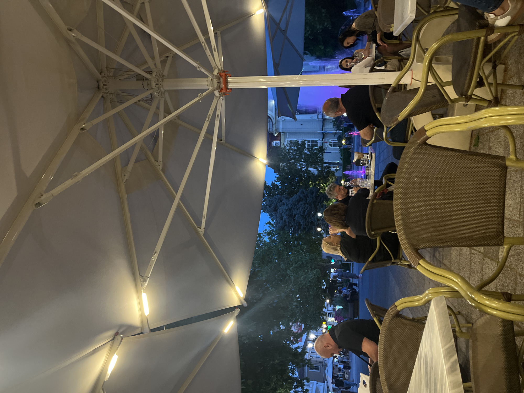 The image depicts an outdoor seating area, likely a restaurant or cafe, in Plovdiv, Bulgaria. An oversized umbrella with integrated lighting dominates the foreground, positioned over tables and chairs. Patrons are seated at the tables, engaged in conversation and dining. In the background, a street scene is visible, with buildings and trees illuminated at dusk. A fountain with colored lights adds to the ambiance. The tables are covered with white marble tops.