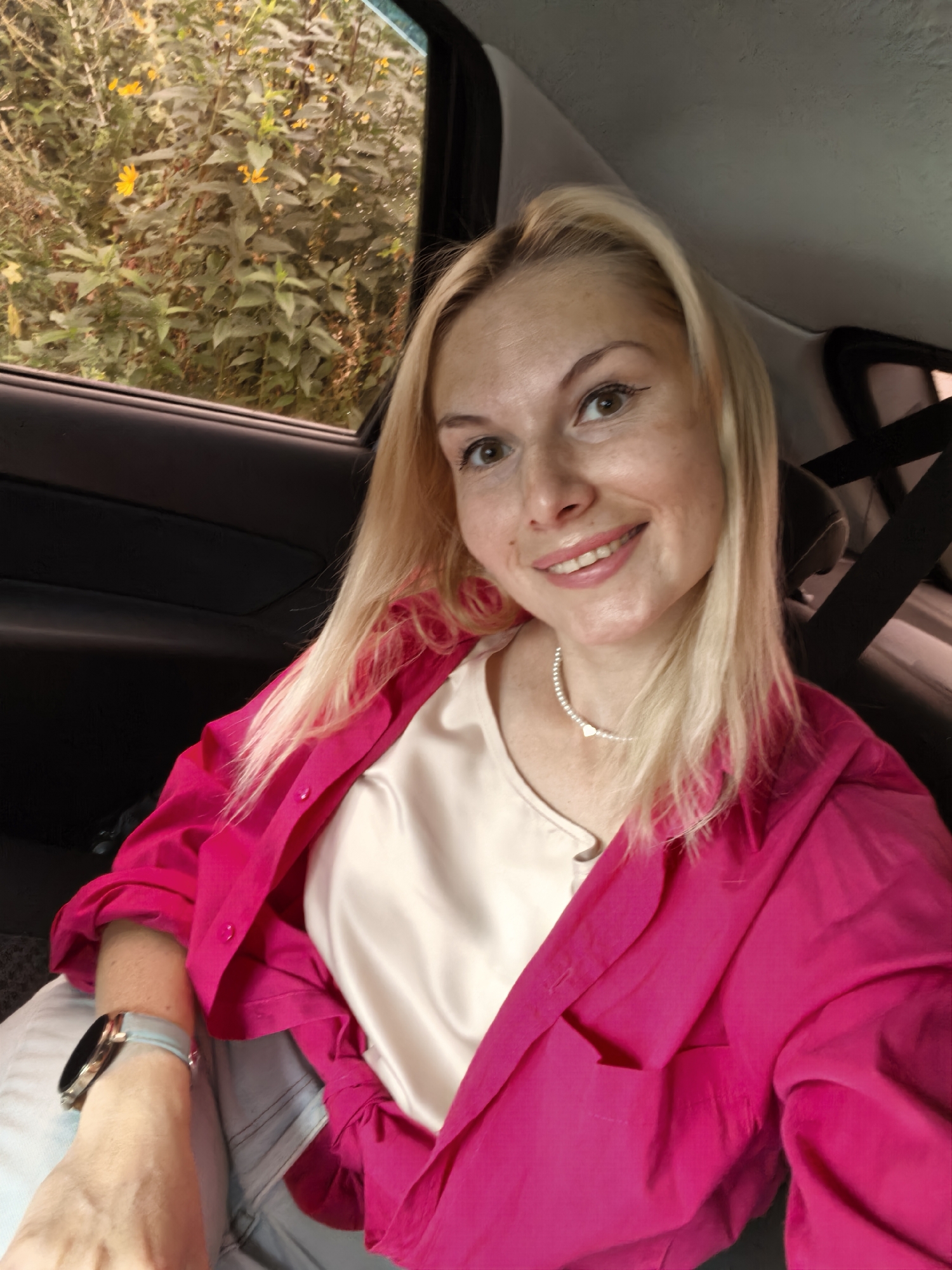 A woman with blonde hair and light skin is taking a selfie from the back seat of a car. She is smiling and looking at the camera. She is wearing a bright pink jacket over a cream-colored top and a pearl necklace. A smartwatch is visible on her left wrist. Through the car window, lush green foliage and yellow flowers are visible. The interior of the car appears to be dark with visible seatbelts. The overall scene suggests a casual outdoor setting, possibly during the daytime. The image does not provide any specific landmarks or indicators that would allow for precise location identification within Sterlitamak, Russia.