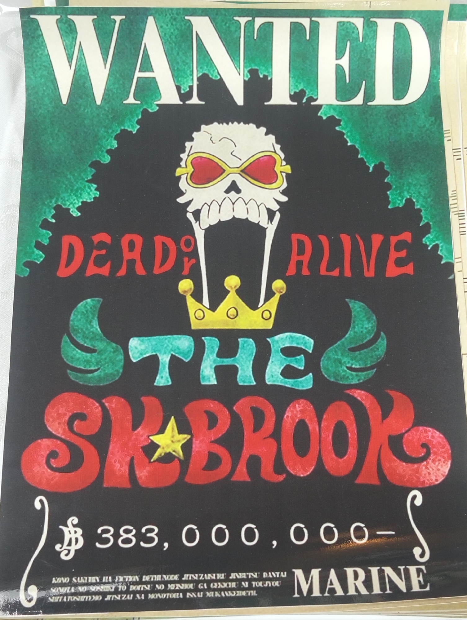 The image is a photograph of a "WANTED" poster with a black background. At the top, the word "WANTED" is displayed in large, white, stylized letters against a green, leafy border. Below this, a skull with heart-shaped sunglasses and a golden crown is depicted. The words "DEAD OR ALIVE" are written in red. In the center, the word "THE" is in teal. Below that, "SK BROOK" is written in large, red, stylized letters with a yellow star on the "A". A dollar symbol followed by "383,000,000" is present at the bottom, with a stylized "S" on the right. In very small text at the bottom, there is Japanese writing and the word "MARINE" in white.
The photograph appears to be taken indoors, likely in a shop or at a convention, given the lighting and the slight angle of the poster. There are no discernible landmarks or features that would indicate a specific location within Dhaka, Bangladesh. The poster itself is the primary subject of the image.