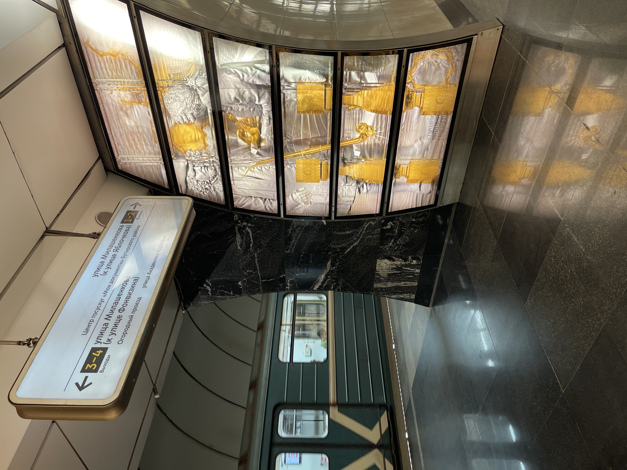 The image shows a metro station platform in an unidentified city, possibly Moscow, based on the Cyrillic script on the signs. A metro train is visible, partially in view along the platform. An illuminated sign indicates exits 3-4 to "улица Милашенкова" (Milashenkova Street) and other points of interest. Above the track, there is an art installation featuring silver and gold-colored figures and objects behind glass panels. The platform floor is made of dark, polished stone, reflecting light and images from the station. The ceilings are reflective. The picture is taken from a slightly elevated perspective looking down at the platform and train.