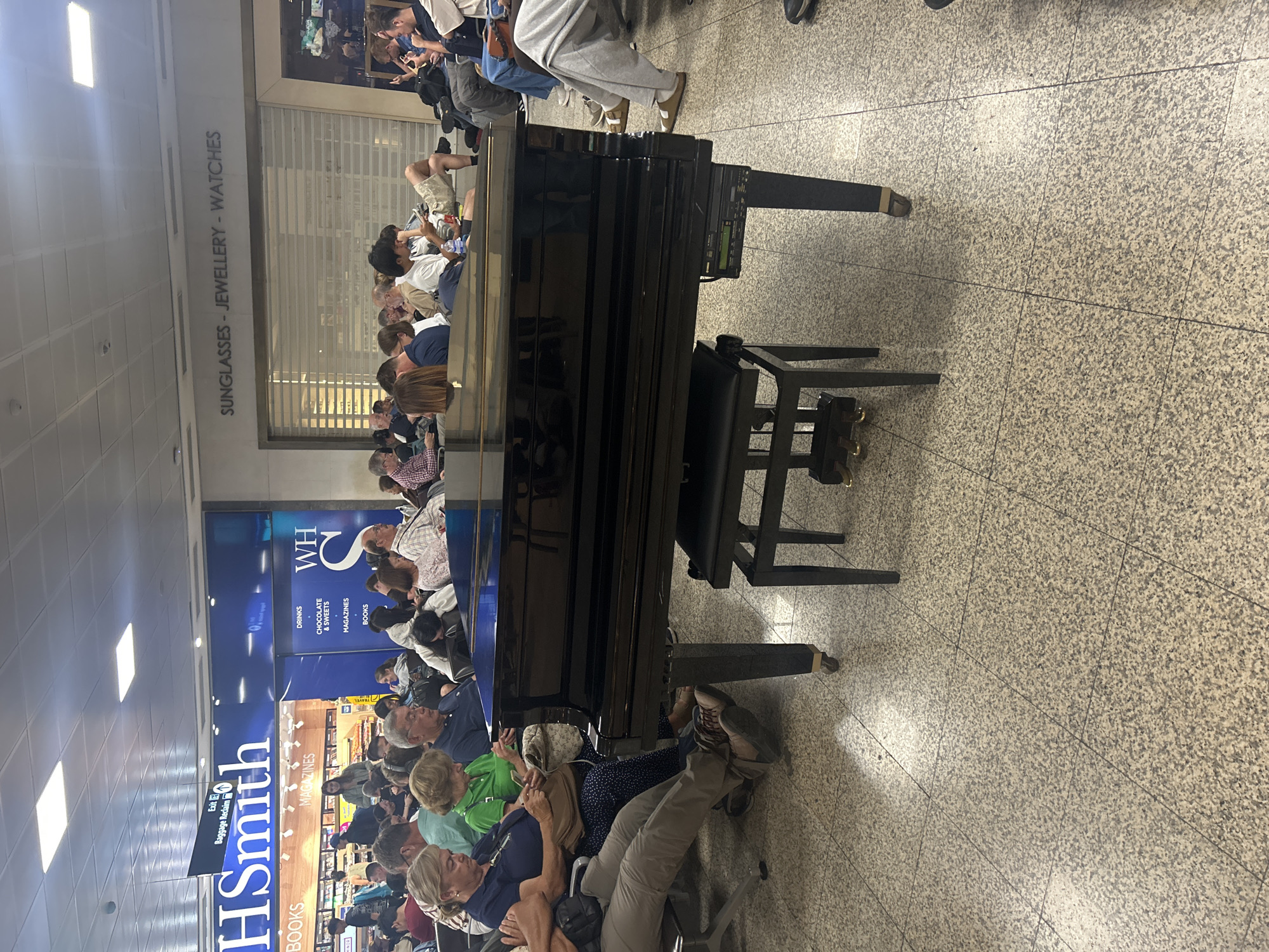 The image shows an airport scene in Luqa, Malta. A black piano is positioned in the foreground amidst a crowd of people, many of whom are seated on chairs. Behind the crowd, signage indicates the presence of a WHSmith store with advertisements for books, magazines, drinks, chocolates, sweets, and travel items. Above the store, a closed storefront displays the words "SUNGLASSES - JEWELLERY - WATCHES". The floor is tiled, and the lighting appears to be typical of an airport terminal. People are waiting around.