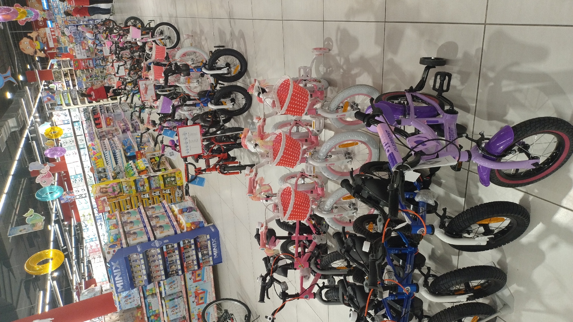 The image shows an interior view of a large retail store, possibly a department store or a toy store, located in Pylaia Municipal Unit, Greece. The foreground and midground are filled with a display of children's bicycles of various sizes and colors, predominantly pink and purple, with some white and blue models visible. Many of the smaller bikes are equipped with training wheels and front baskets, some decorated with ribbons.
In the background, shelves stocked with toys and other merchandise are visible. There are also some adult shoppers and store employees in red shirts browsing the aisles or standing near the shelves. The environment is well-lit with overhead fluorescent lighting. Decorative elements like colorful inflatable rings and cartoon character cutouts are hanging from the ceiling, suggesting a festive or child-oriented atmosphere. The floor is tiled in a light color. The overall impression is a well-organized retail space dedicated to children's products.