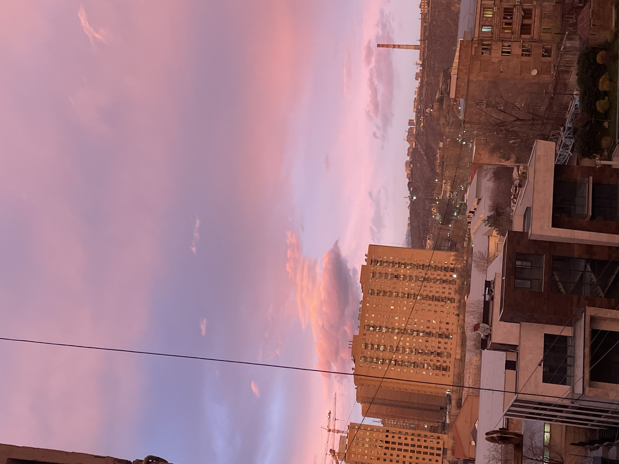 The image depicts an urban landscape in Yerevan, Armenia, under a vibrant pink and purple twilight sky, likely at dusk. A dark, thin utility cable vertically bisects the left side of the frame. In the midground, several large, multi-story residential or commercial buildings constructed of light brown material exhibit numerous illuminated windows. To the far left, two prominent orange tower cranes indicate ongoing construction. Below these, a dense array of varied smaller buildings, including modern structures with dark glass and older, light-colored buildings with white roofs, extends towards the foreground. Bare trees are interspersed among the structures. In the distant background, a large hill or mountain is covered with scattered city lights and additional buildings, crowned by a tall, red and white striped industrial chimney on the far right horizon. No individuals, specific actions, or direct interactions are discernible within the scene.