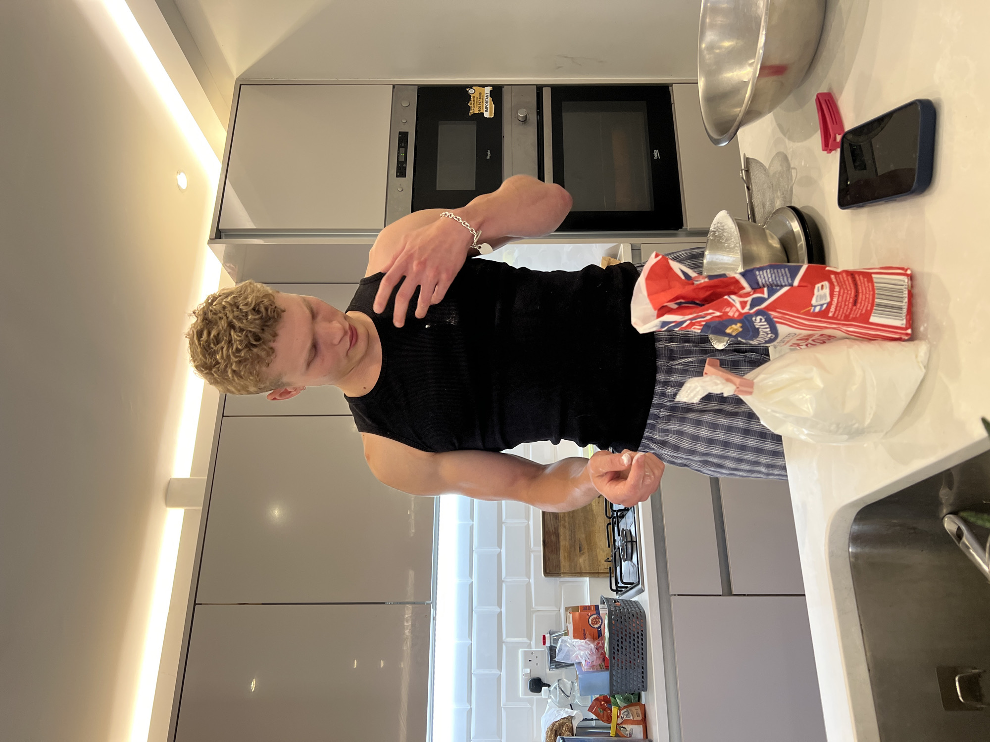 A young man with curly blonde hair and a black tank top is standing in a modern kitchen, likely in London, United Kingdom. He is wearing plaid pajama bottoms and is in the process of baking. In front of him, a bag of flour is open and spills onto the counter. A scale and a bowl are also present, indicating measurements are being taken. The kitchen features sleek grey cabinets and white subway tile backsplash, with ovens visible in the background. A phone and a red clip are on the counter, along with a large metal bowl. The lighting in the kitchen is warm and bright, with under-cabinet lighting visible.