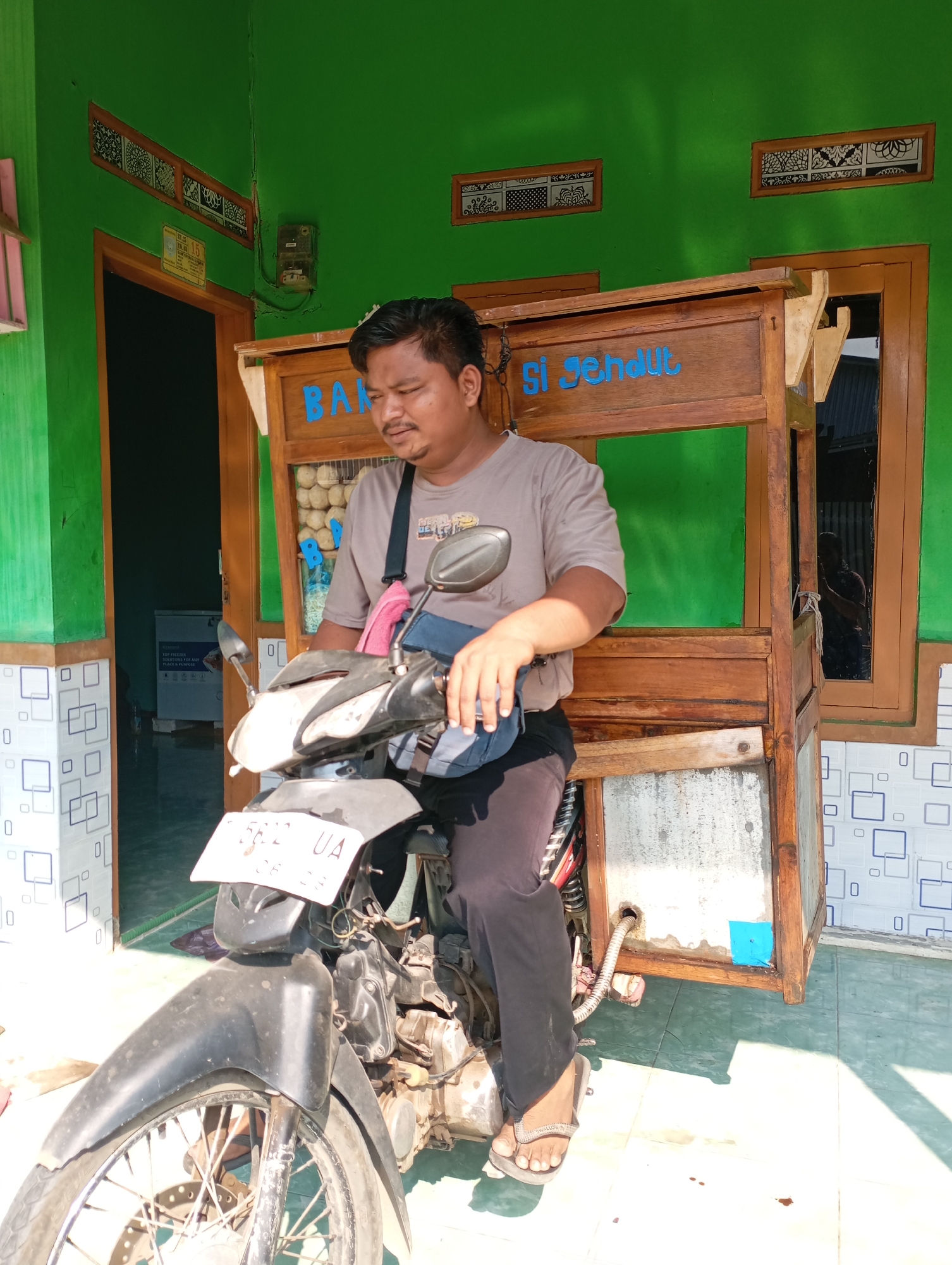 A man sits on a motorcycle in front of a food stall, likely in Kecamatan Haurgeulis, Indonesia. The stall, made of wood, has the words "BAK SICHENDUT" painted in blue on its upper section. Inside the stall, spherical food items are visible. The man is wearing a grey t-shirt, dark pants, and flip-flops. He has a bag slung over his shoulder. The motorcycle is black and has a license plate that reads "5672 UA". The background features a bright green wall with decorative window elements and tiled sections. A doorway with a wooden frame is also visible to the left, leading into a dimly lit interior. The ground is tiled, and there are scattered dark spots, possibly oil stains.