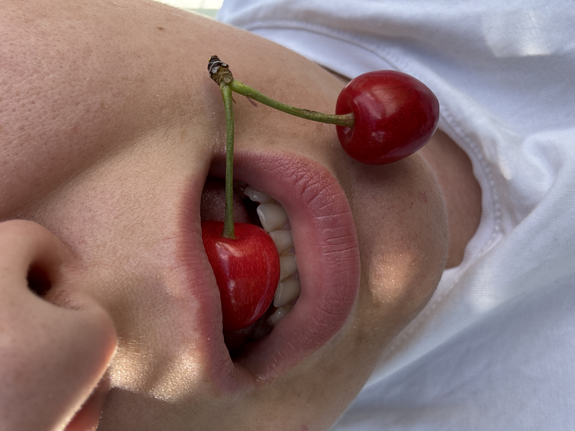 A close-up, waist-up shot shows a person wearing a white t-shirt, with two cherries near their mouth. One cherry is held between the person's teeth, while the other hangs from a stem on the right side of their face. The person's nose and lips are visible, as well as some teeth. The background is blurred and out of focus, showing a hint of a light-colored surface. The lighting suggests an outdoor setting, possibly during the day, with sunlight casting shadows and highlights. The image is devoid of any specific indicators of the city of Добровское сельское поселение, Ukraine.