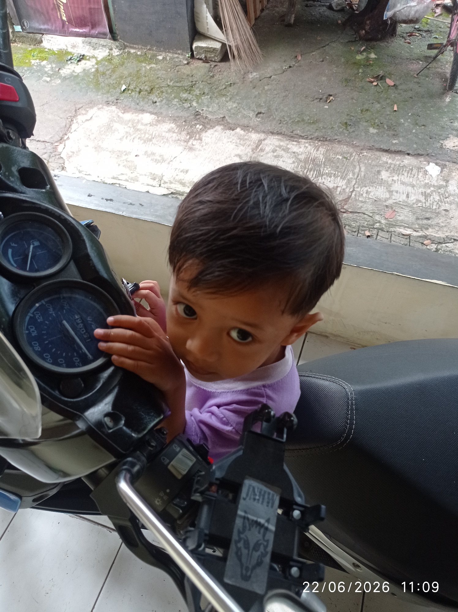 A young child, appearing to be under the age of five, is seated on a motorcycle, looking towards the camera. The child has dark hair and is wearing a purple shirt. They are positioned between the motorcycle's handlebars and the seat. The motorcycle's dashboard, with visible gauges, is to the left of the child. The child's hands are near the dashboard. The background shows a cracked concrete surface with some debris, suggesting an outdoor urban environment. A portion of a building and some foliage are visible in the upper background. A date and time stamp "22/06/2026 11:09" is present in the lower right corner of the image. This scene is located in the city of Sukaraja, Indonesia.