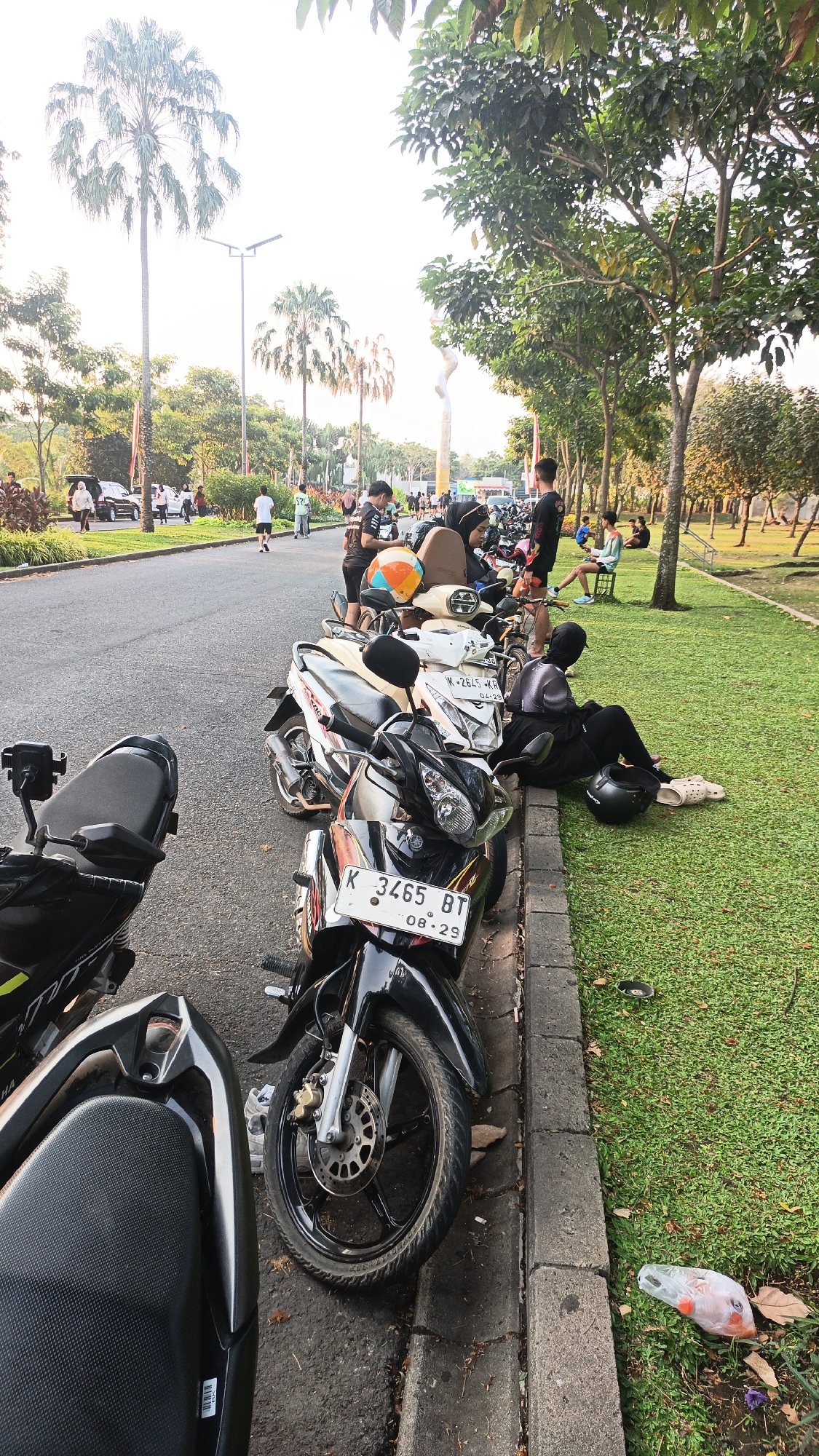In the city of Bacin, Indonesia, a street scene unfolds with a prominent line of parked motorcycles along a curb. The motorcycles are predominantly scooters, with several individuals gathered around them. One person is seated on the grass next to a motorcycle, wearing dark clothing and a headscarf, with a helmet resting nearby. Another individual is leaning against a scooter, holding an orange and blue helmet. In the background, palm trees and lush green grass indicate an outdoor recreational area or park. People are seen walking and standing along a paved pathway adjacent to the road, with cars also visible in the distance. The overall ambiance suggests a casual gathering or a stop during an outing. The presence of motorcycles suggests a mode of transportation common in the area. The lighting indicates it is likely late afternoon or early morning.