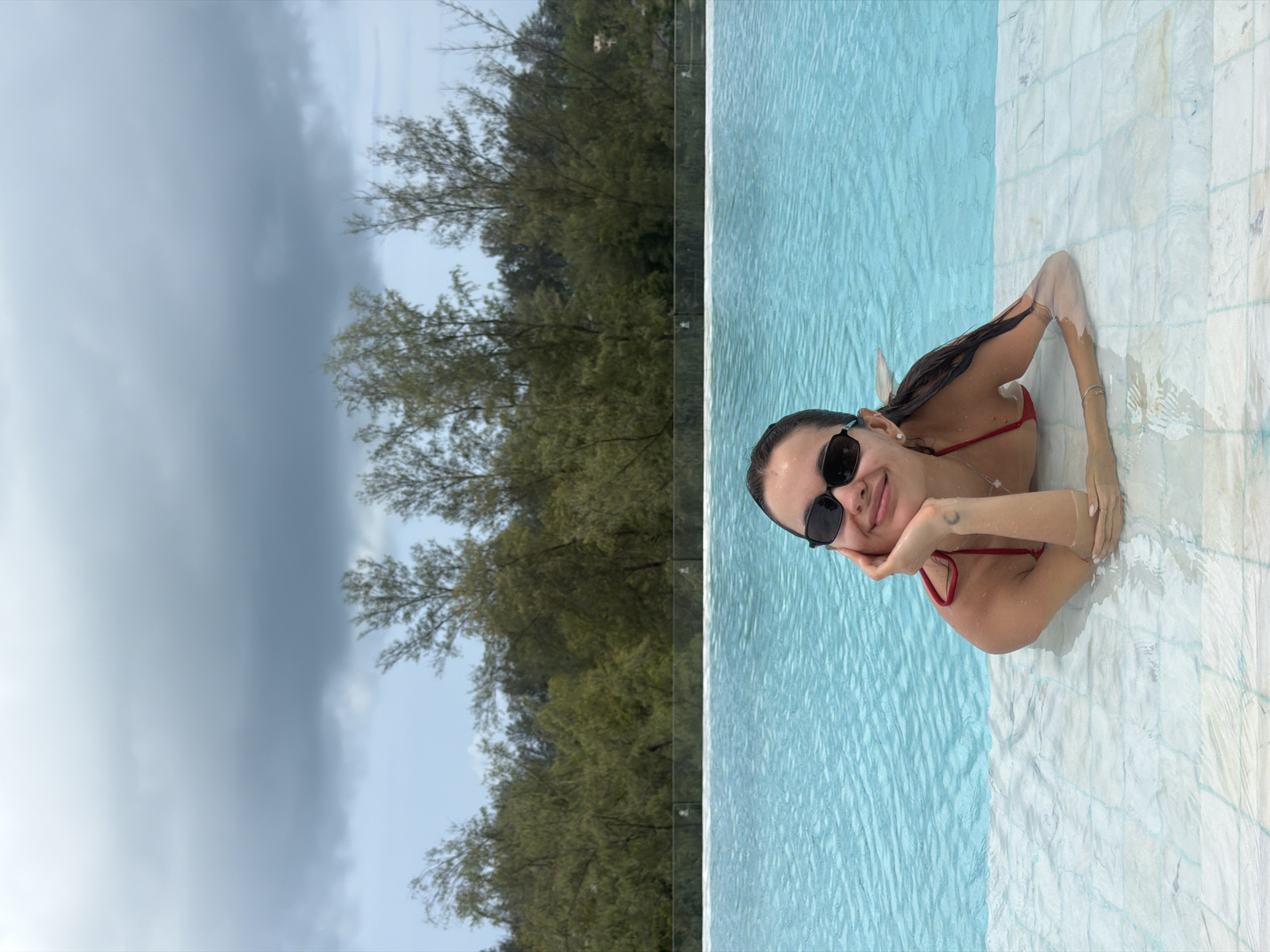 A woman is in a pool at an elevated vantage point. She is wearing a red swimsuit and sunglasses. Her left hand is holding her chin as she is smiling toward the camera. The pool’s edge is made of textured, light-colored tiles. Outside of the pool is a dense area of green trees. The sky above the trees is overcast with light and dark grey clouds. This location is in Choeng Thale, Thailand.