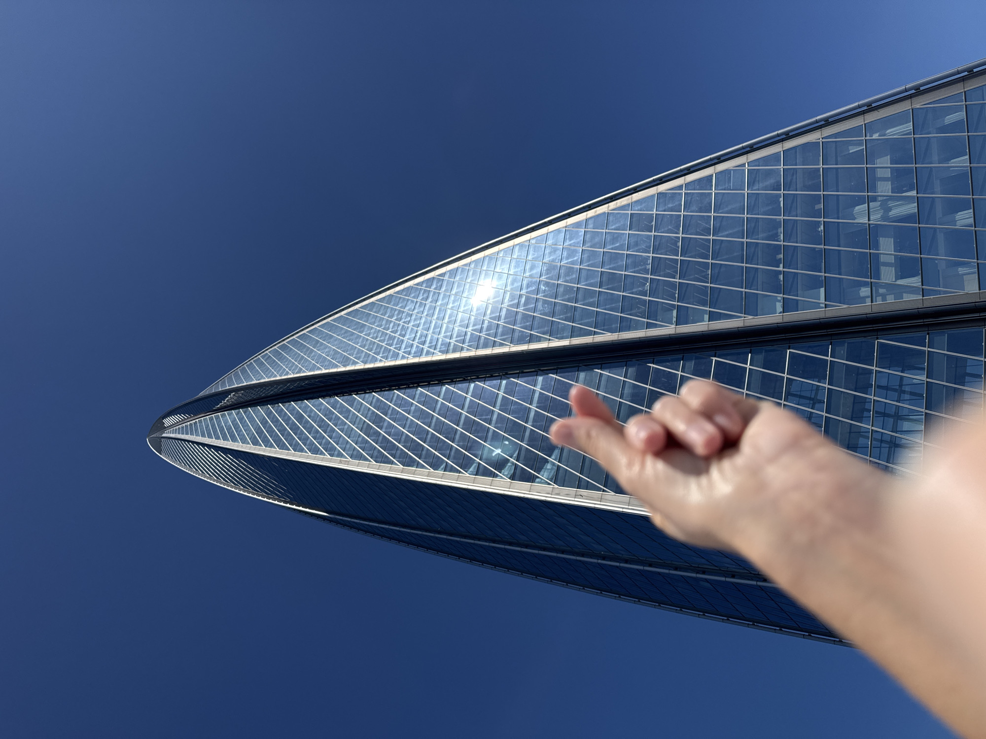An upward-facing, low-angle photograph captures a modern glass skyscraper against a clear, deep blue sky. In the foreground, slightly out of focus, is a person's right hand, angled as if reaching towards or playfully interacting with the building. The hand forms a gesture where the thumb and index finger touch, commonly known as a "finger heart."
The prominent subject in the background is a contemporary glass skyscraper, identifiable as the Lakhta Center in Saint Petersburg, Russia, given its unique twisted and tapering design. Its facade is composed of numerous reflective glass panels, which shimmer brightly under the sun. A strong sun glare is visible reflecting off the glass surface, indicating bright daylight. The sky is a uniform, intense blue with no clouds, suggesting perfectly clear and sunny weather. The perspective emphasizes the immense height and architectural complexity of the building, juxtaposed with the human hand in a whimsical manner. No visible text is present in the image.
