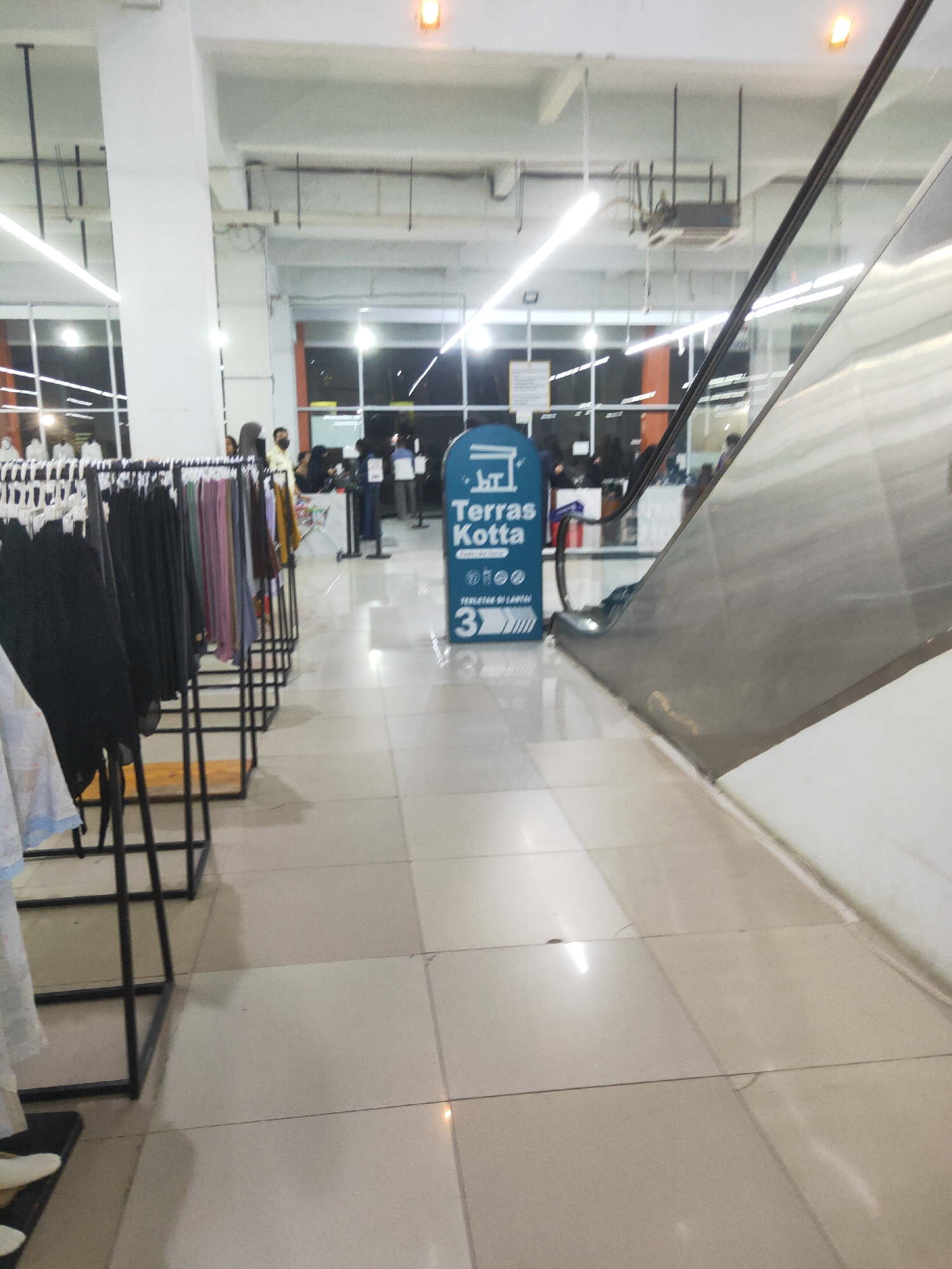 This media file depicts an indoor scene, likely a retail environment such as a department store or shopping mall, in Sintang, Indonesia. The foreground is dominated by clothing racks displaying various garments, primarily in dark and muted colors, with some lighter colored items visible on the left. The floor is tiled with light-colored squares.
In the middle ground, an escalator is visible, its metallic surface reflecting the overhead lighting. To the left of the escalator, a blue sign stands upright, featuring text in Indonesian. The text reads "Terras Kotta" at the top, followed by a symbol resembling a building and then more text. Below this, there are icons and the number "3" with arrows pointing to the right, suggesting a floor number or direction. People are scattered throughout the background, some appearing to be shoppers browsing the clothing racks, while others are near the escalator and the sign. Some individuals are wearing masks. The overall lighting suggests it is daytime, but the indoor setting makes it difficult to ascertain external weather conditions. The atmosphere appears to be one of normal commercial activity.