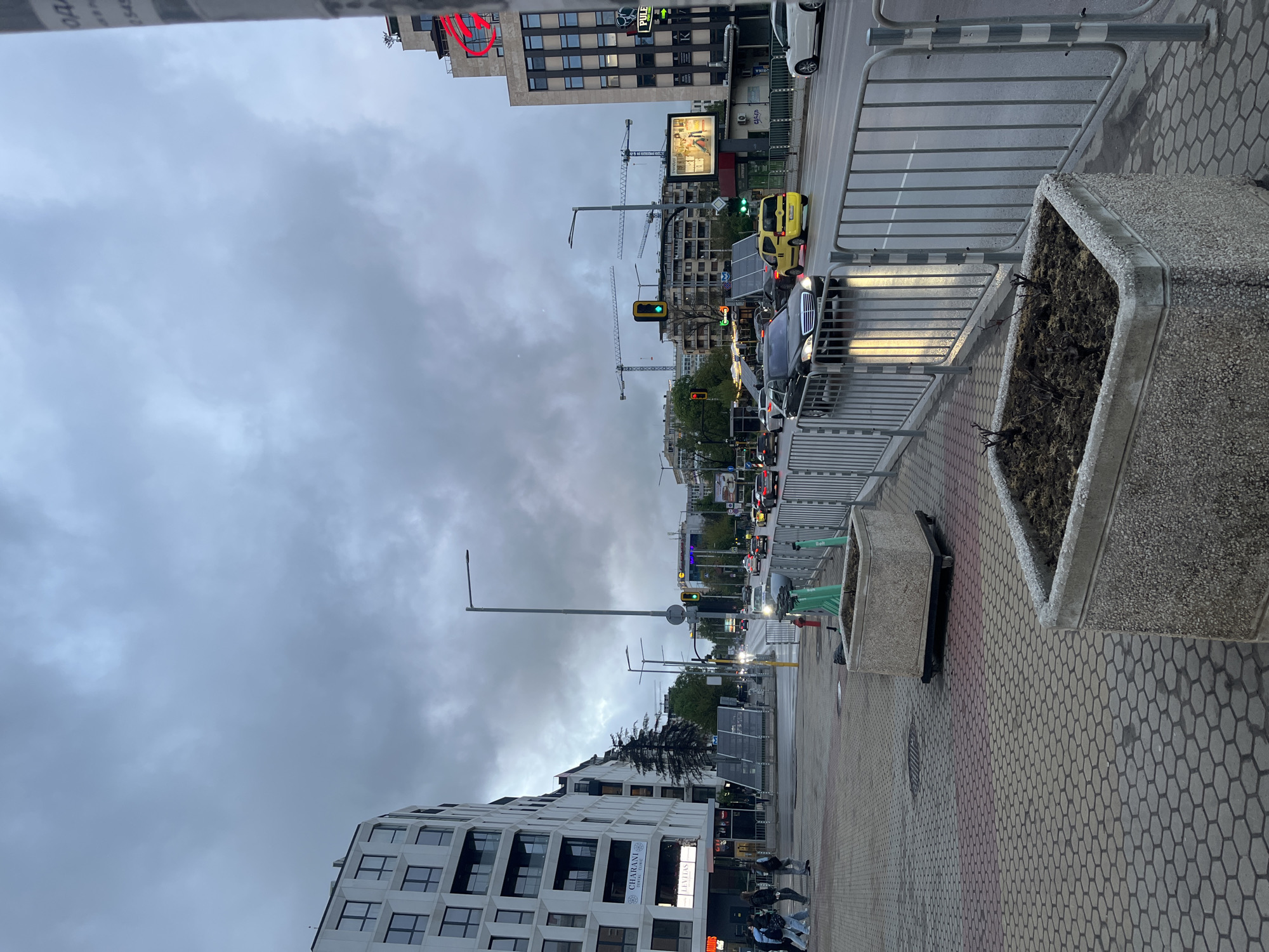An urban street scene in Sofia, Bulgaria, captured on an overcast day, likely during late afternoon or early evening. The image is oriented sideways, showing a street with active traffic, a sidewalk, and multi-story buildings under a grey, cloudy sky.
**Subjects:** The scene includes several vehicles on the road, such as a distinctive yellow car (possibly a taxi) and other dark-colored cars, some with headlights illuminated. A small group of people are visible walking on the sidewalk in the foreground. Construction cranes dot the skyline in the background, indicating ongoing development.
**Objects:** The street features traffic lights, street lamps, and metal barriers separating the road from the sidewalk. The sidewalk is paved with hexagonal blocks in shades of grey and a reddish strip. Large concrete planters, one filled with dark soil and sparse, dry-looking plants, are present along the sidewalk. Green electric scooters, identifiable as "Bolt" brand, are parked on the sidewalk, alongside a red fire hydrant. Buildings on both sides of the street exhibit modern architecture; one on the left has prominent geometric window patterns. An illuminated billboard and other signs are visible on the buildings.
**Activity/Event:** The scene depicts typical city life with vehicular traffic, pedestrians, and ongoing construction. Cars appear to be moving or waiting at a traffic light, suggesting a routine urban flow.
**Notable Details:** The sky is heavily clouded, giving a muted, slightly damp atmosphere. The road surface appears wet, possibly from recent rain. Car headlights are on, reinforcing the impression of low light or poor visibility due to weather.
**Visible Text:** Signs on buildings display "PULĘ", "CHARANI STYLE & BEAUTY", and "LEVIES". "Bolt" is visible on the green scooters. A partially visible red neon sign on a building shows Cyrillic characters, appearing to be "ОБЪ".