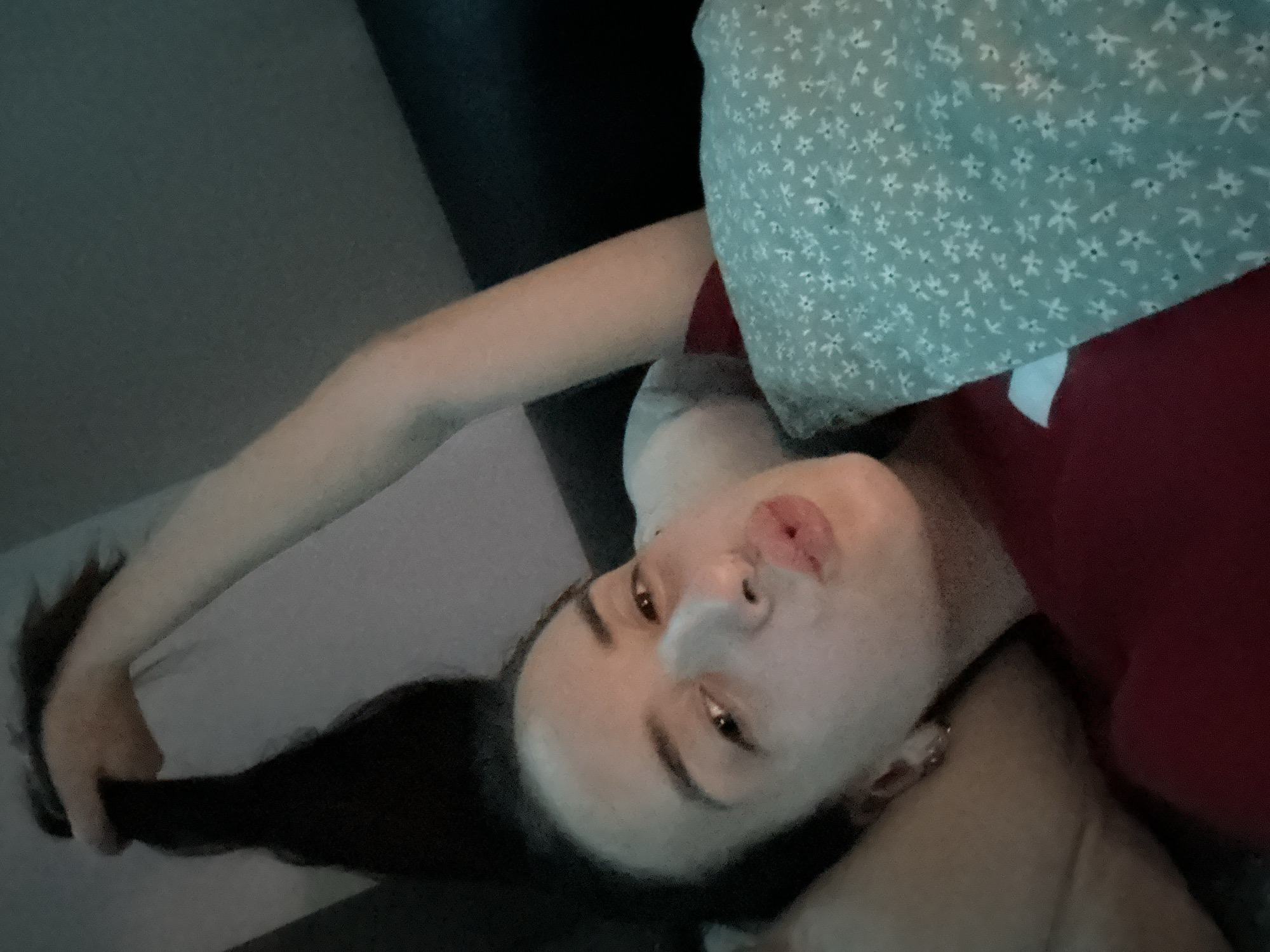 A young woman is lying on her side, holding a lock of her dark hair up with her right hand. She is wearing a red shirt and a light blue pillow with a floral pattern is visible beside her. Her left arm is resting on a pillow. Her face is turned towards the camera, and she has her lips pursed as if for a kiss. The background is dark and appears to be an indoor setting, possibly a bedroom. The image was taken in Shumen, Bulgaria.