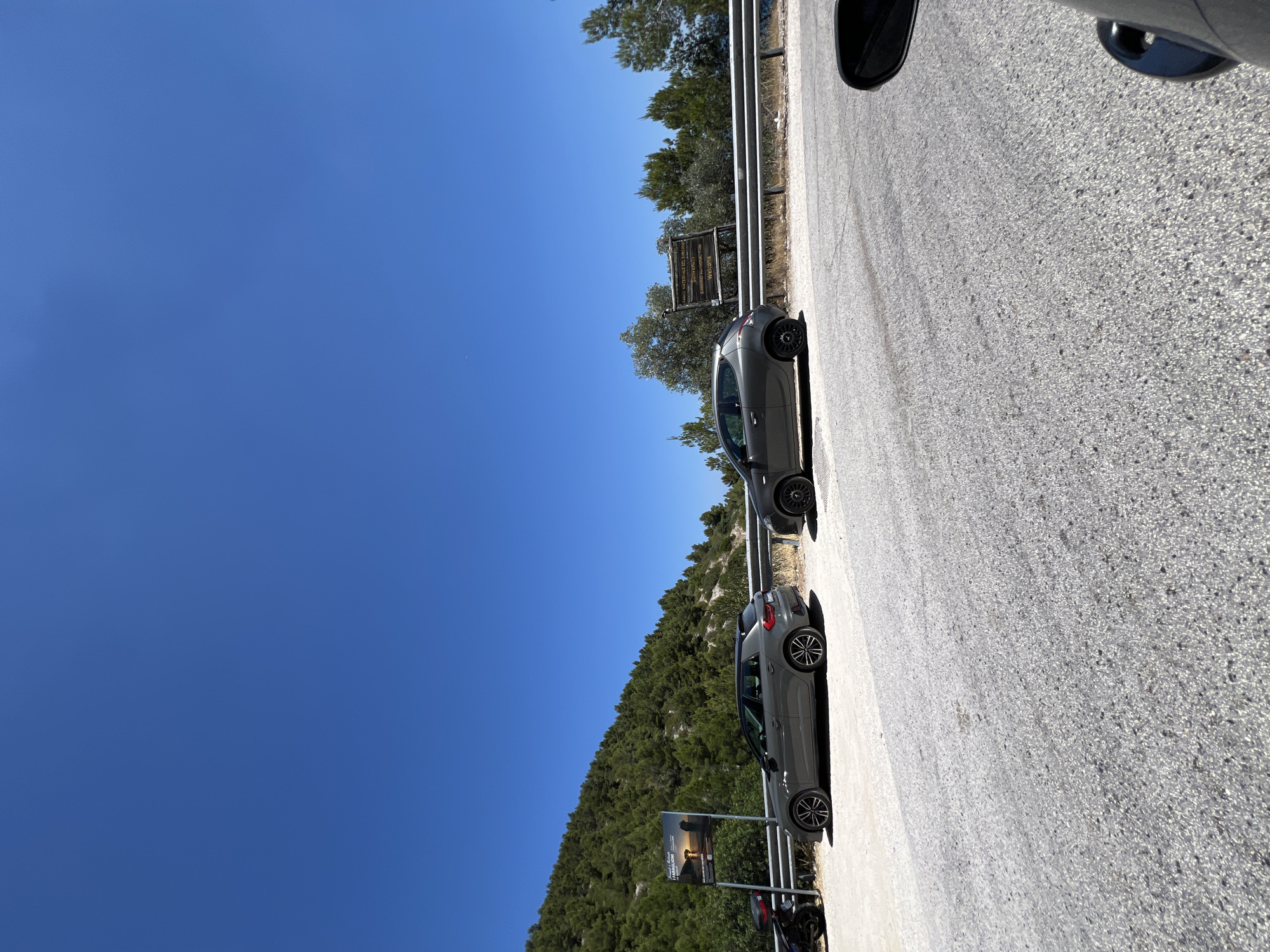 On a sunny day, two gray cars are parked on the shoulder of a road that curves along a densely wooded hillside. The road surface is asphalt with visible gravel. To the left, a dark-colored motorcycle is parked next to a sign displaying an image of rock formations and text that reads "Faraglioni." Further down the road, to the right, another sign is visible with text in Italian and English, indicating "Parco Nazionale del Gargano" and "Welcome." The hillside is covered in a thick growth of green trees, and the sky is a clear, vibrant blue. The scene appears to be in or near Mattinata, Italy, given the mention of the Gargano National Park and the Faraglioni rock formations, which are a notable feature of the Mattinata coastline.