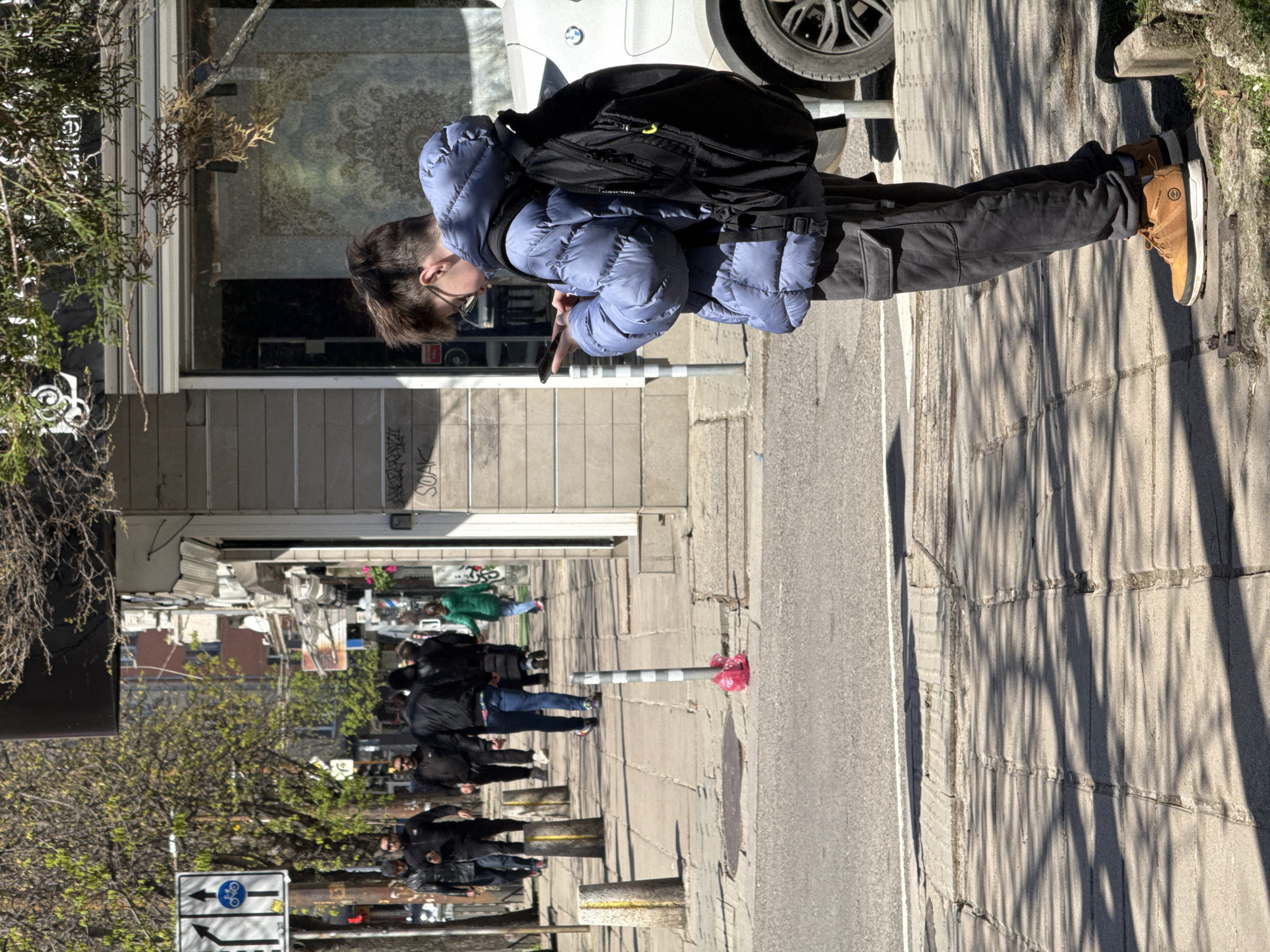 The image captures a street scene, possibly in Sofia, Bulgaria. A person wearing a blue puffy jacket, dark pants, and brown work boots stands on the sidewalk, looking at their mobile phone. They have a black backpack on. The sidewalk is paved with rectangular stones and casts long shadows. Cars are parked nearby, and a few pedestrians walk along the sidewalk in the background. The buildings along the street are various styles, with shops on the ground floor and apartments above. A road sign and a bollard wrapped in a red plastic bag are visible near the pedestrian's path.