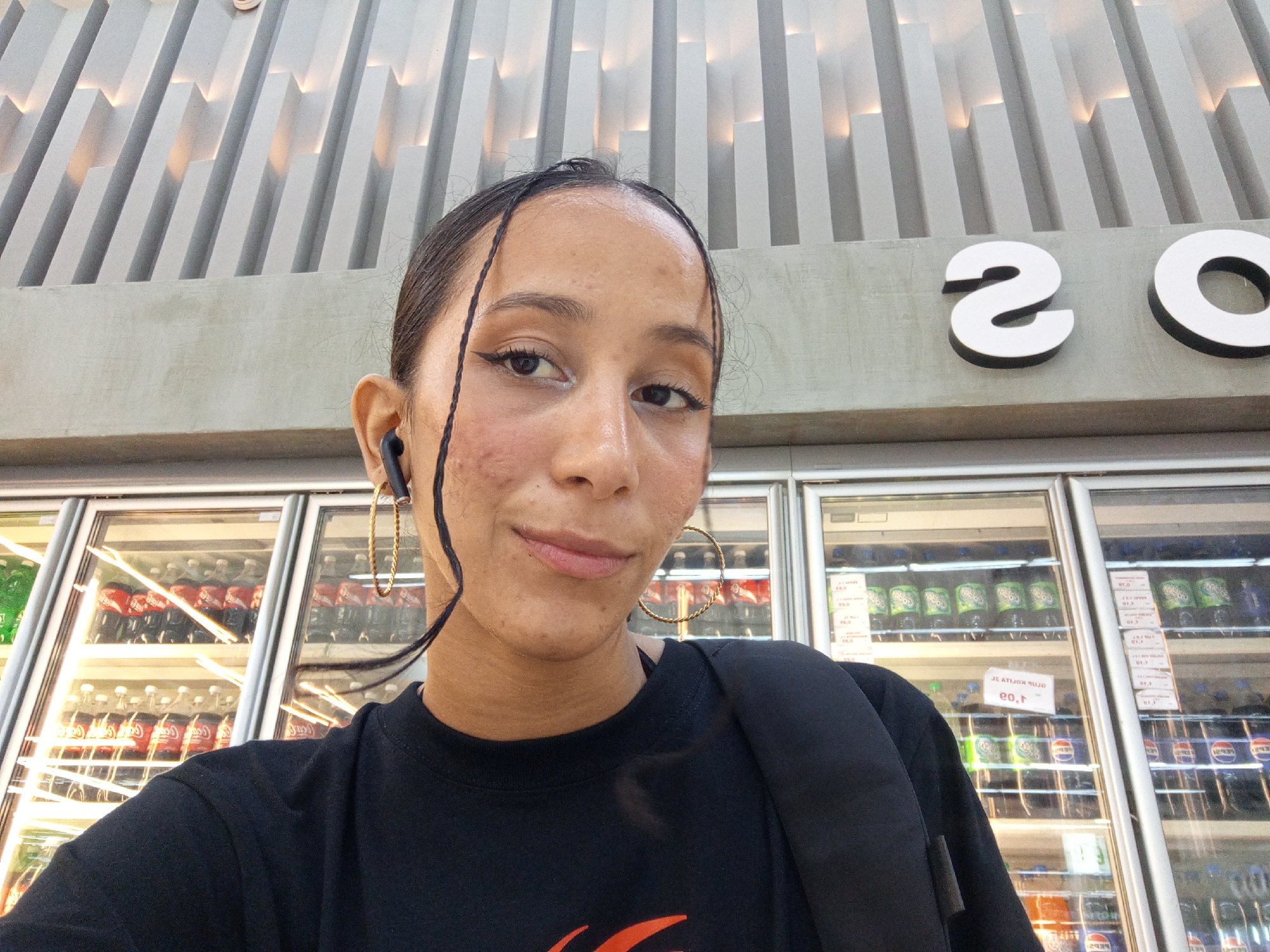 A woman with dark hair pulled back, wearing gold hoop earrings and black wireless earbuds, is taking a selfie. She is wearing a black t-shirt and carrying a black backpack. She stands in front of refrigerators filled with soda bottles and other beverages. Above the refrigerators, there is a sign that says "20". The ceiling features a modern design with white, angled slats. The image was taken indoors, presumably at a market in Porlamar, Venezuela.